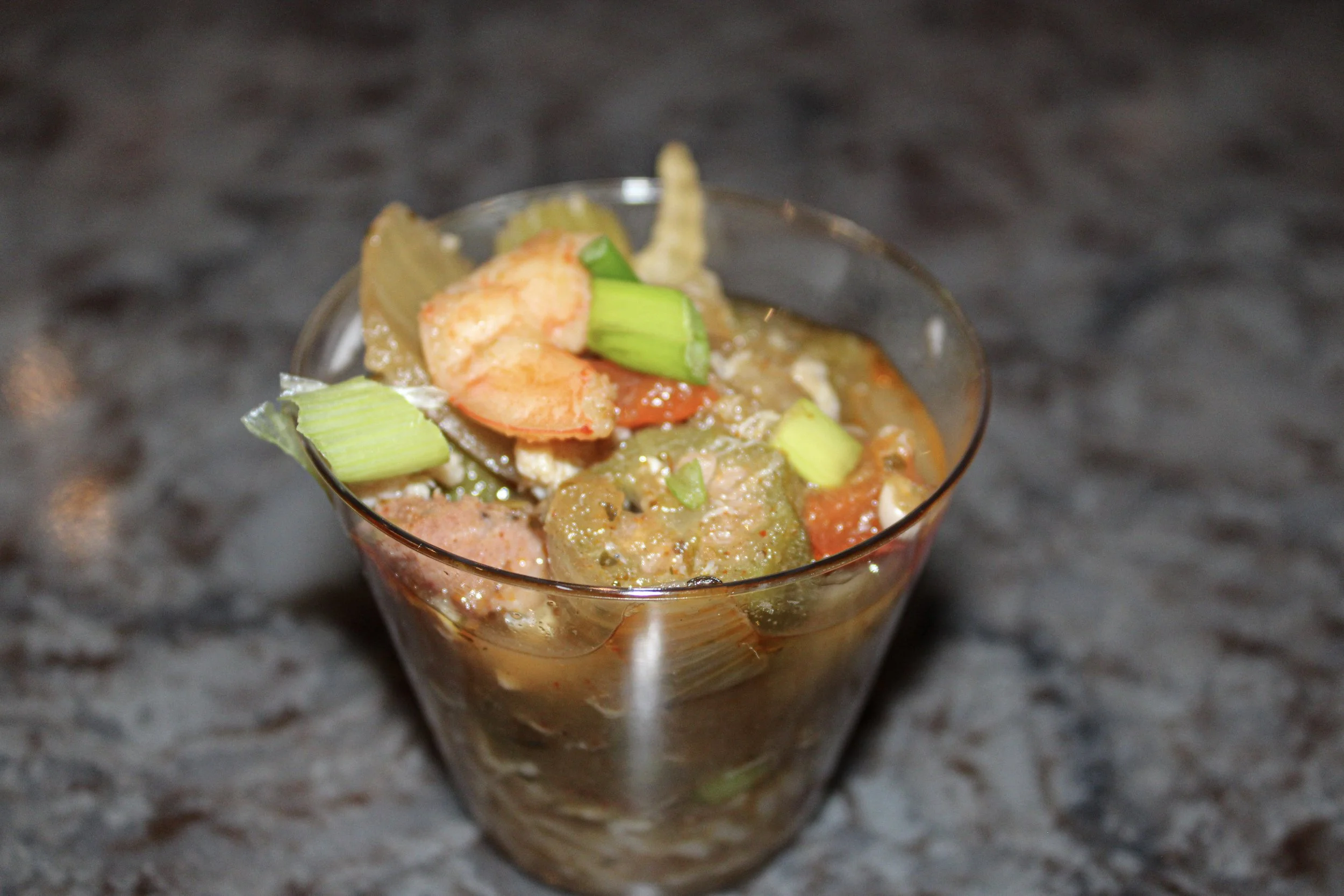 A close-up of a shrimp and vegetable dish served in a clear glass, with celery and other vegetables visible, set against a dark, textured surface.