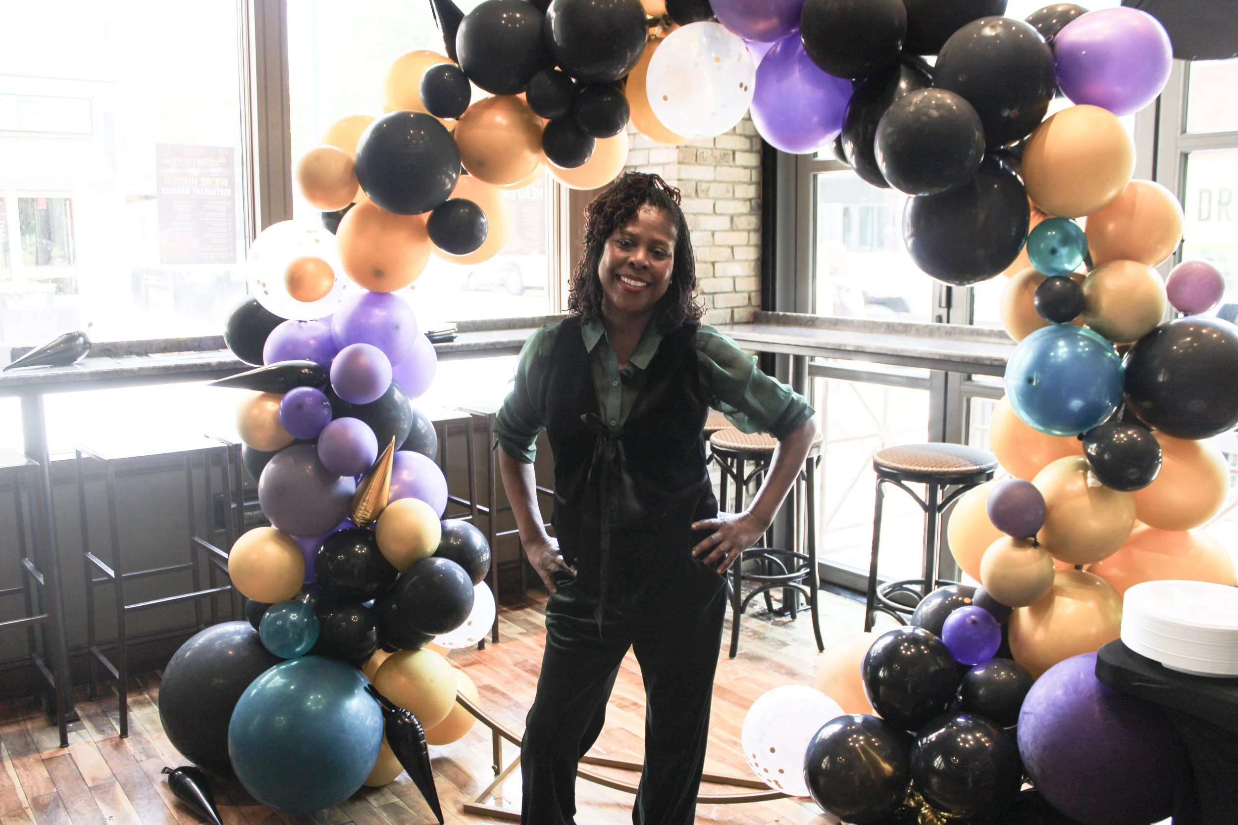 Woman standing inside a room with large balloon arch in black, purple, gold, and teal colors, smiling at the camera.