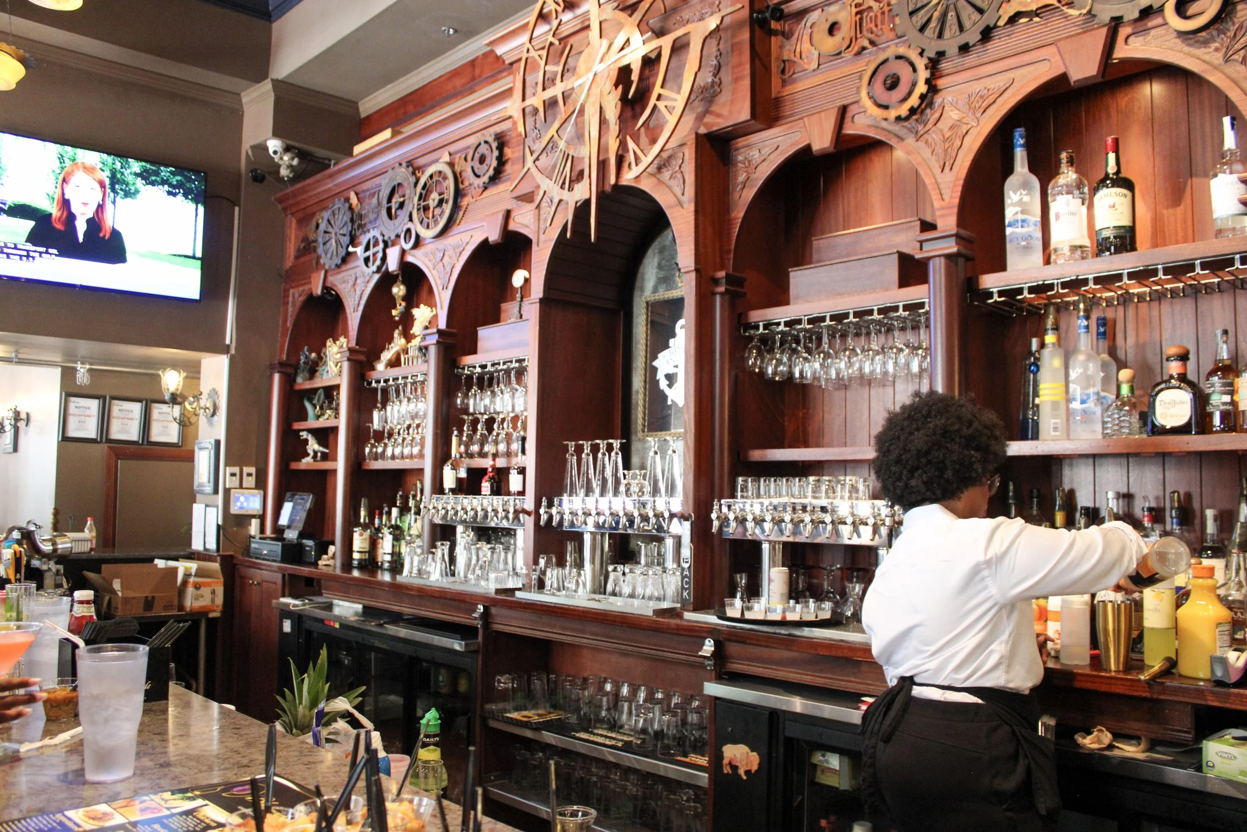 A bartender in a white shirt and black apron working behind a wooden bar with liquor bottles and glassware in a restaurant or bar setting. A large decorative wooden wall with gears and clock details is above the bar, and a television screen is visible in the upper left corner.