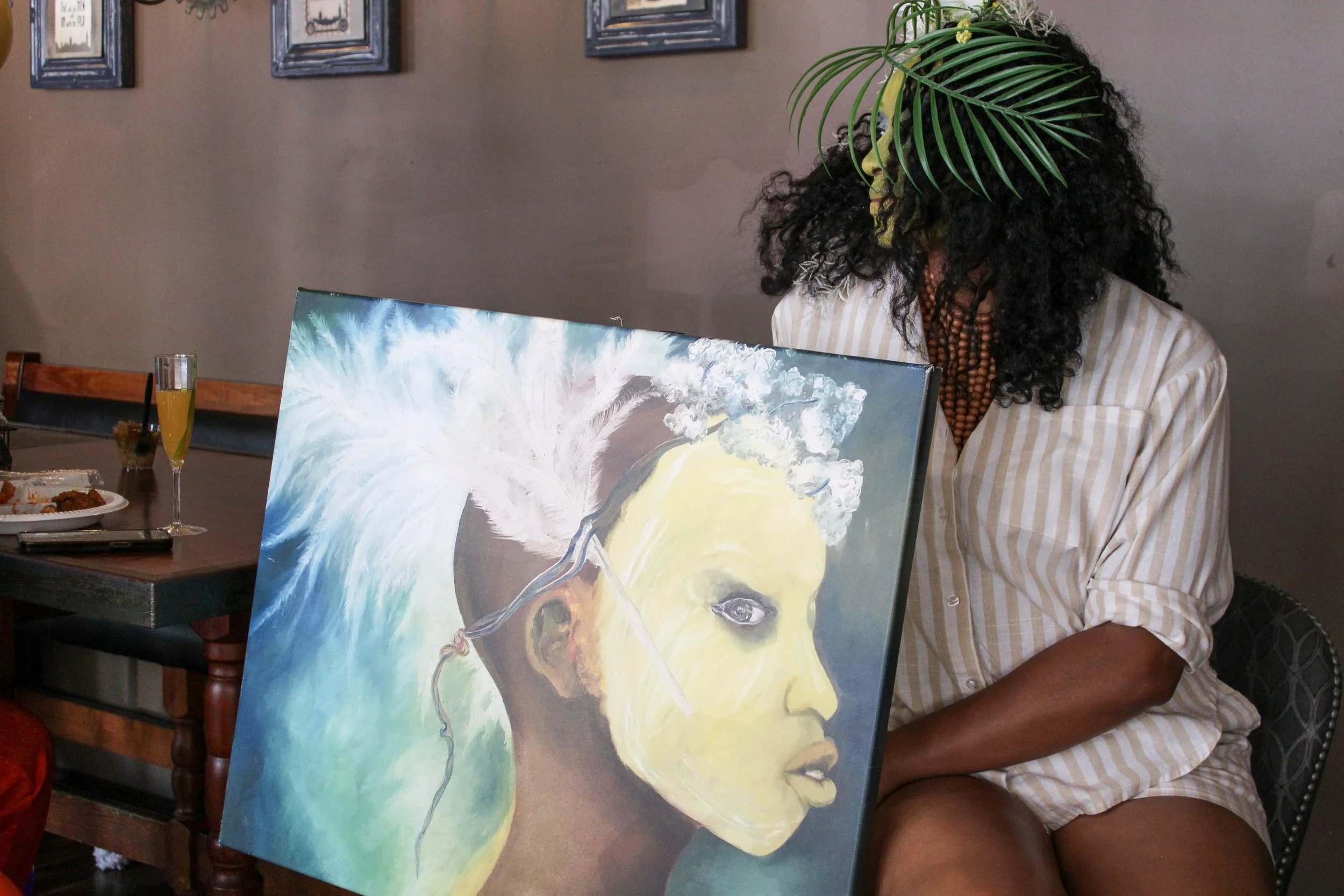 A woman with curly black hair wearing a striped shirt is sitting indoors, partially obscured by a colorful painting of a woman's face with abstract elements. There are framed pictures on the wall, and a table with food and a glass of orange juice are visible in the background.