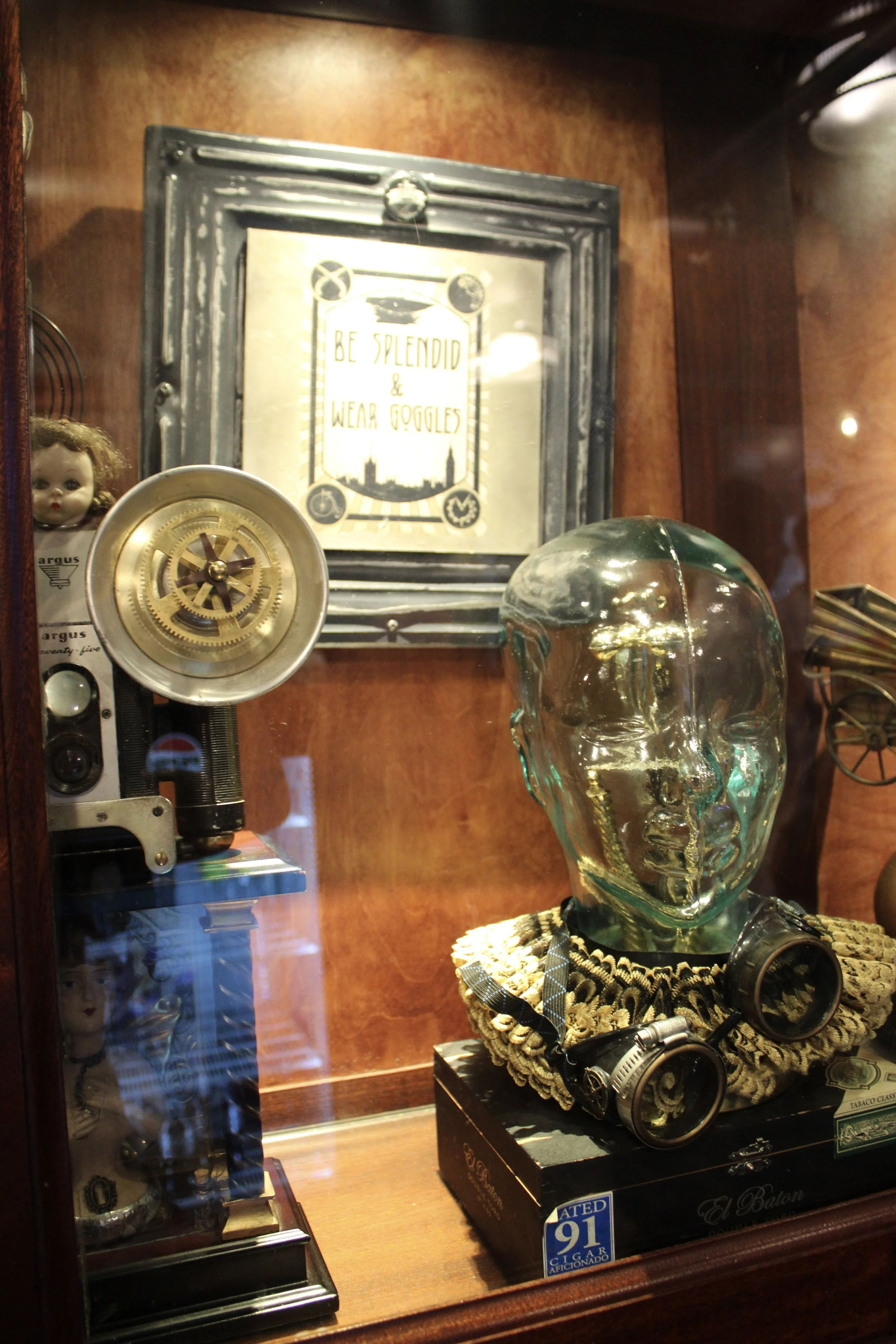 A display of various objects, including a glass skull, a vintage-style lamp, a doll, a sign that says 'Be splendid & wear goggles,' and some vintage camera equipment.