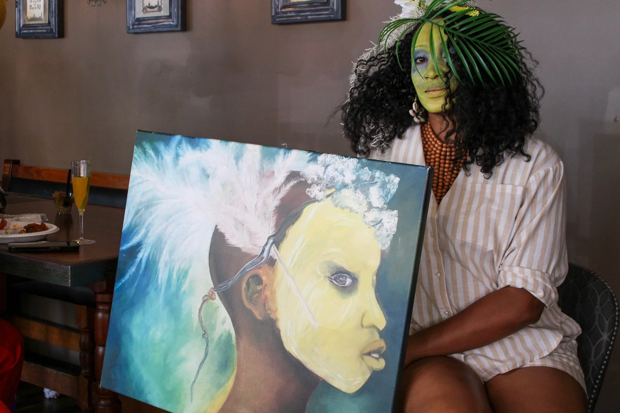 A woman with dark curly hair, painted green face, wearing colorful jewelry, holding a painting of a woman's face with a splash of blue and white background, sitting at a table in a room with framed pictures on the wall.