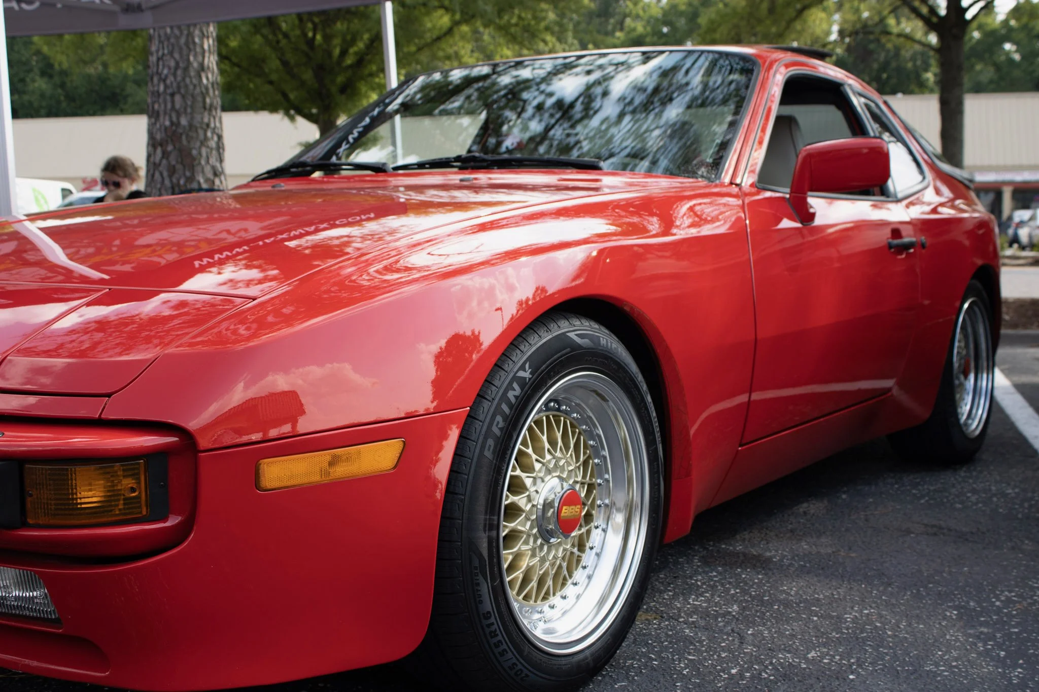 Close-up of a shiny red sports car with gold alloy wheels parked outdoors, with trees and a person in the background.