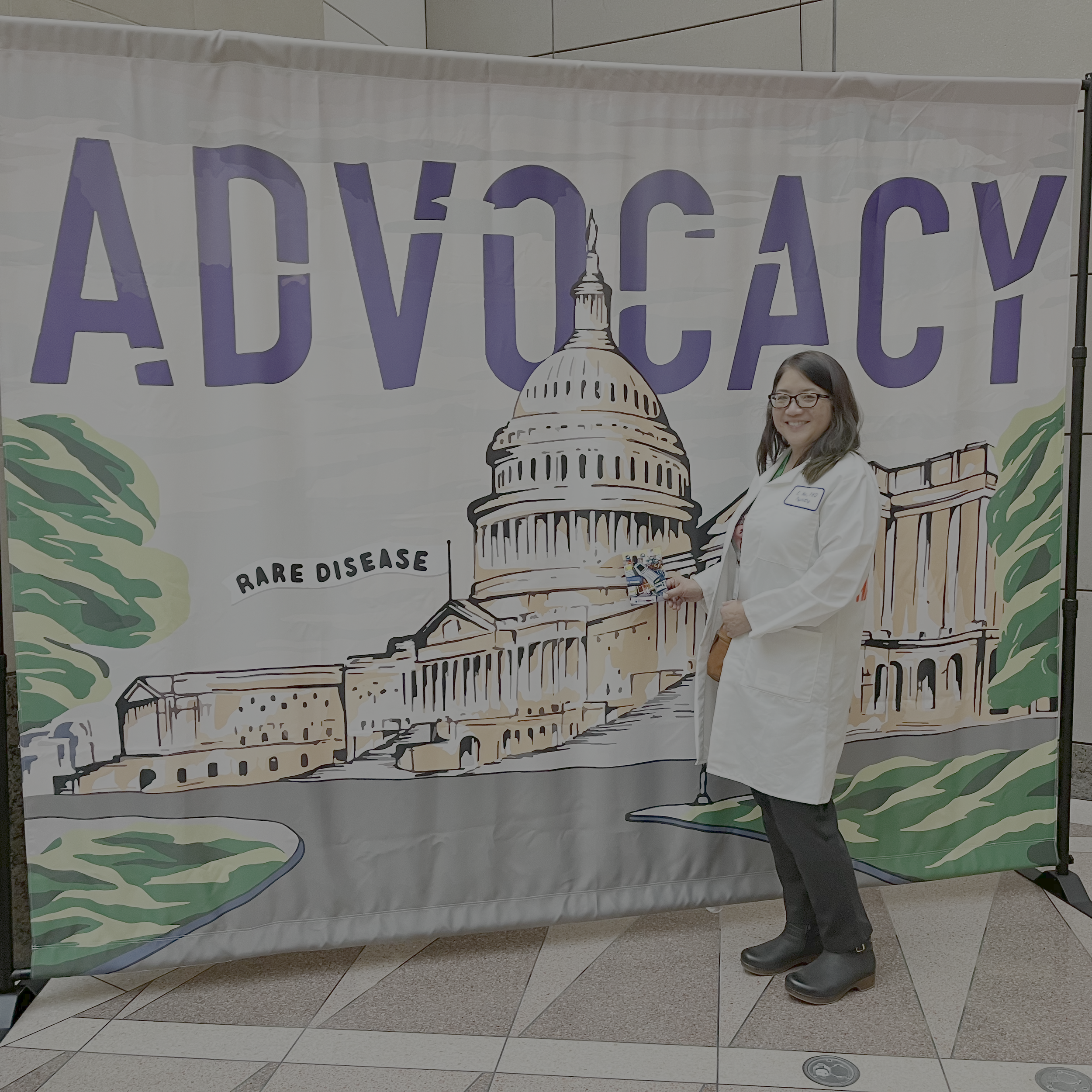 Person standing in front of a large banner displaying the Capitol building with the words 'RARE DISEASE' and the word 'ADVOCACY' written prominently, along with part of the phrase 'FOR.' The person is wearing glasses, a white coat, and black shoes, holding a pamphlet or brochure.