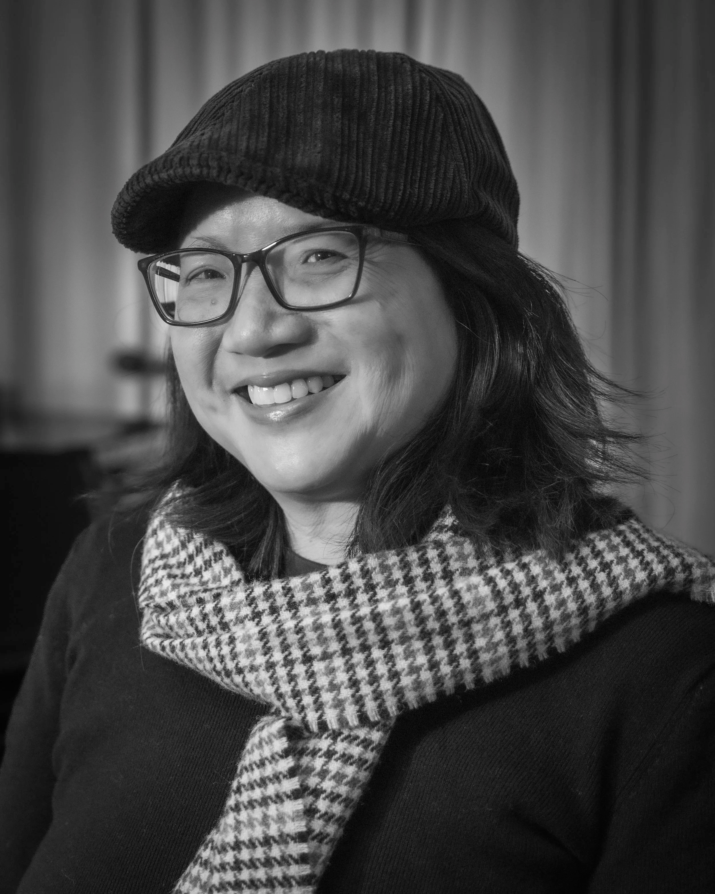 A smiling woman with glasses wearing a dark beret and a houndstooth patterned scarf, with a blurred curtain in the background.