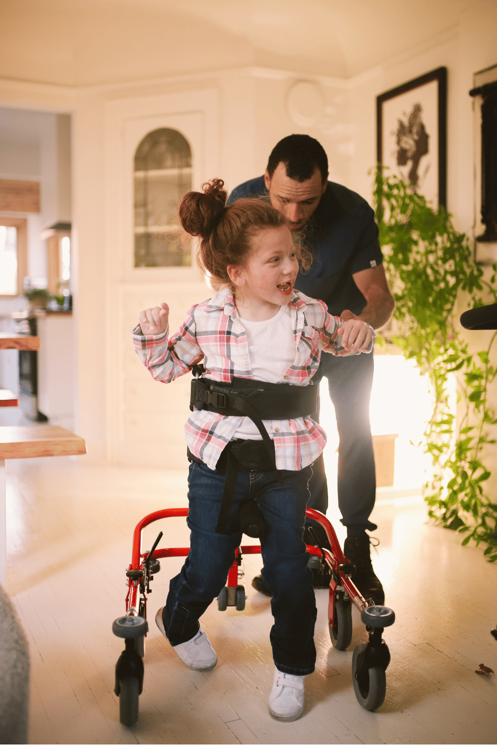 Une jeune fille utilisant une marchette avec l'aide d'un adulte dans une maison lumineuse.