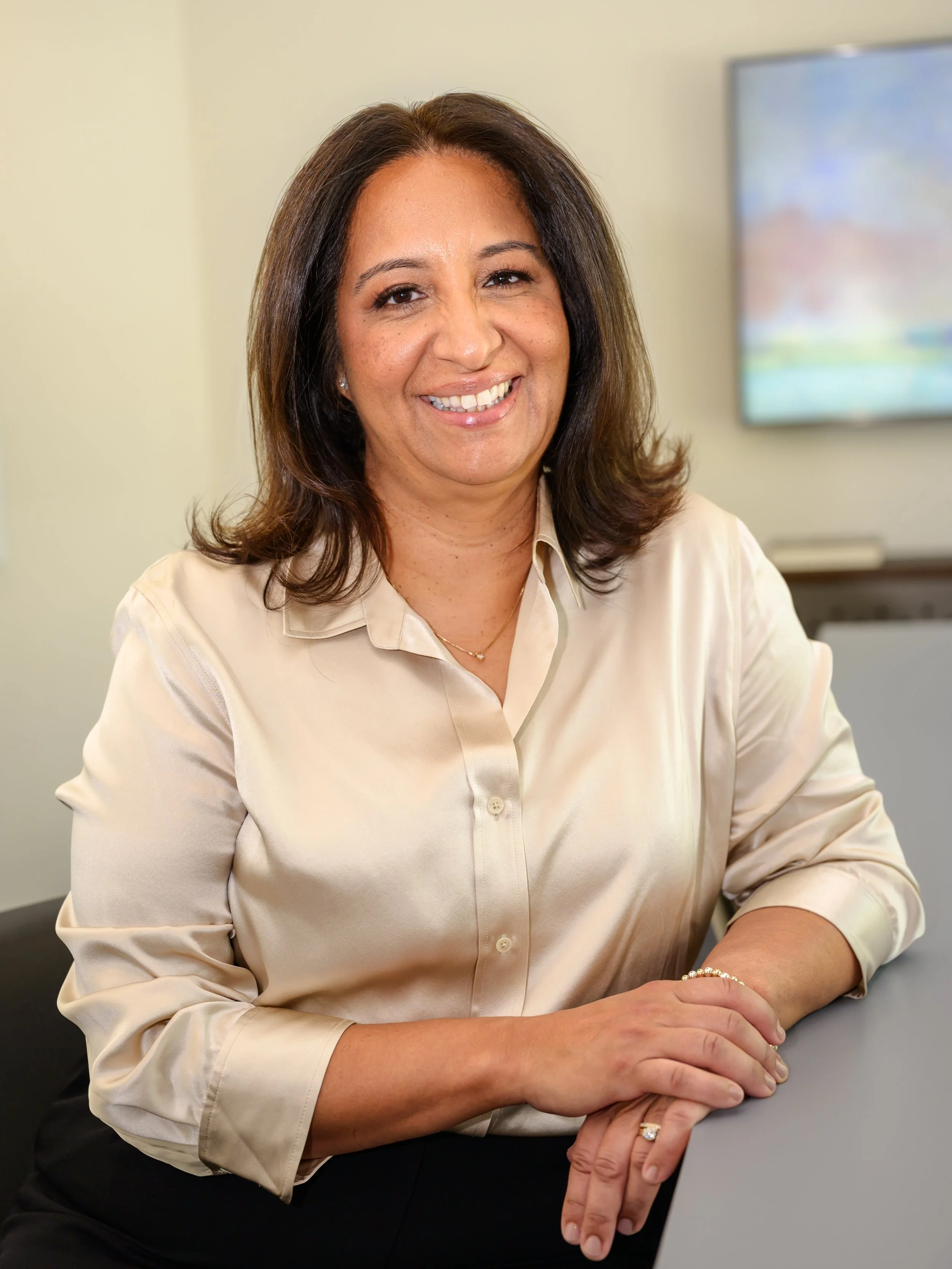A woman with shoulder-length brown hair, wearing a cream-colored blouse, smiling and sitting at a desk with her hands resting on the surface. There is a monitor in the background.