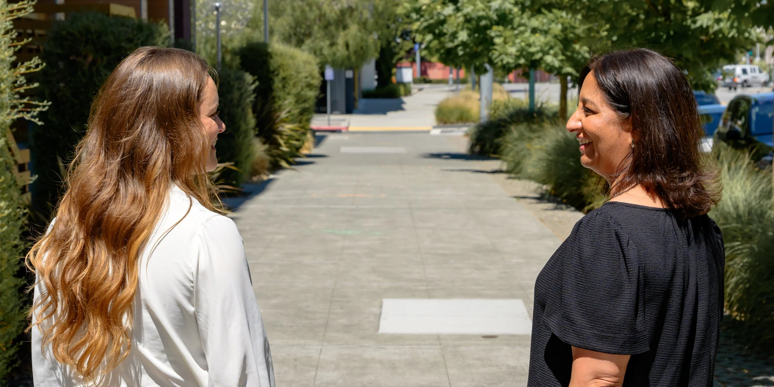 Two women standing face to face on a sidewalk, smiling at each other with trees and parked cars in the background.