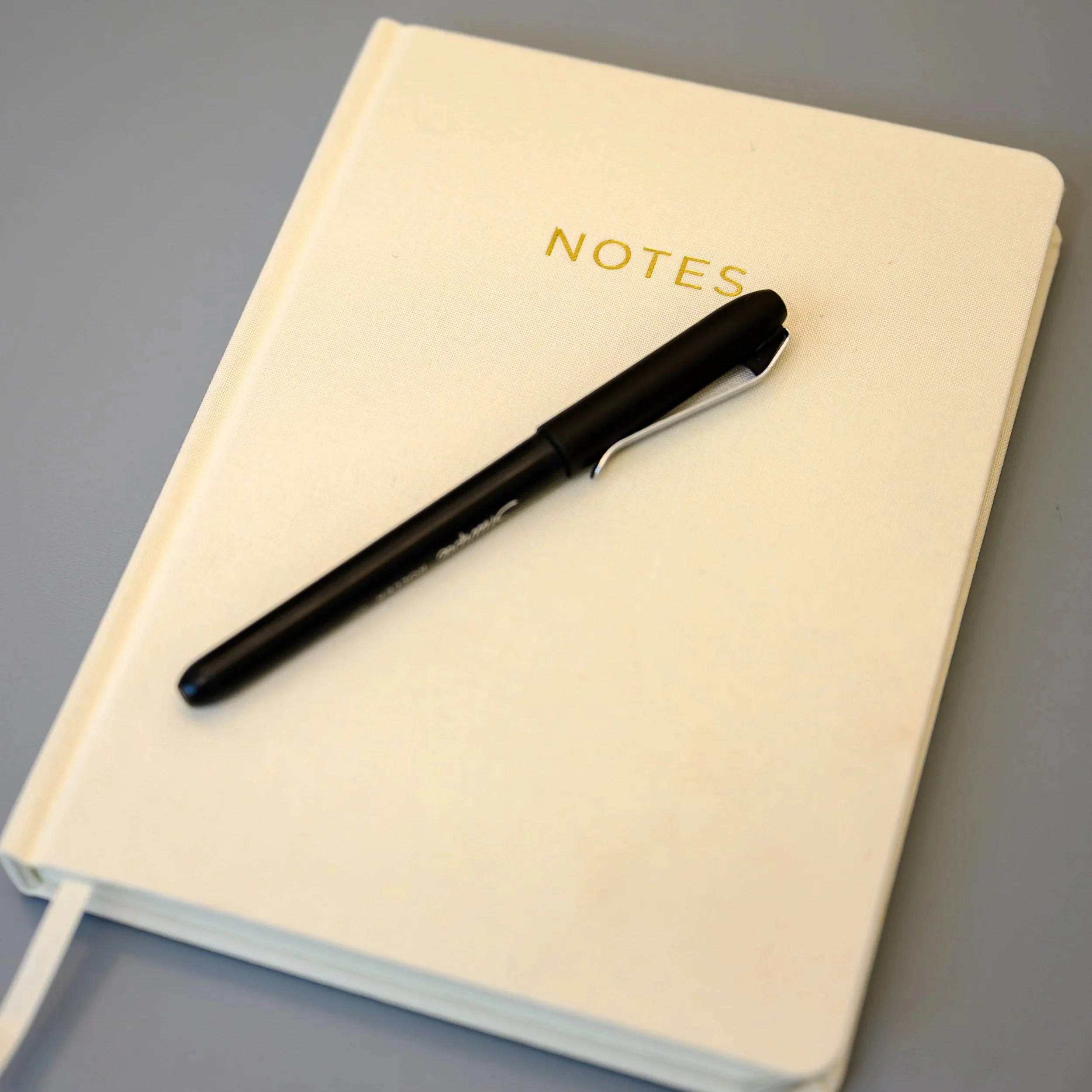 Close-up of a cream-colored notebook with the word 'NOTES' embossed in gold lettering on the cover. A black pen rests on top of the notebook. The background appears to be a gray surface.