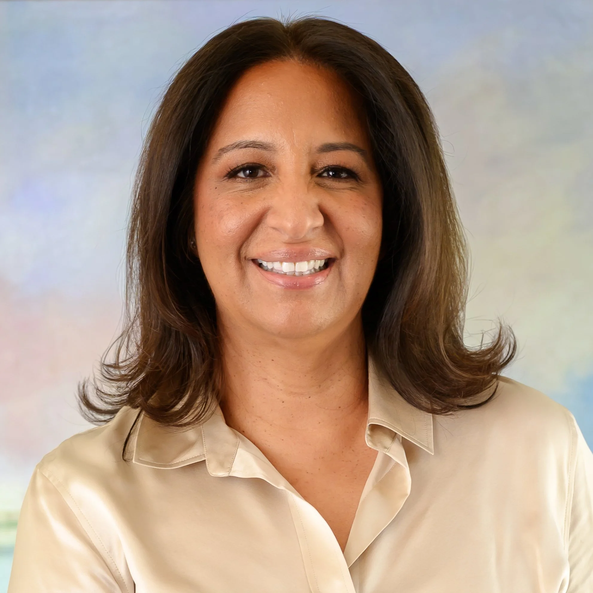 Close-up of a smiling woman with shoulder-length brown hair, wearing a beige collared shirt, against a blurred light background.