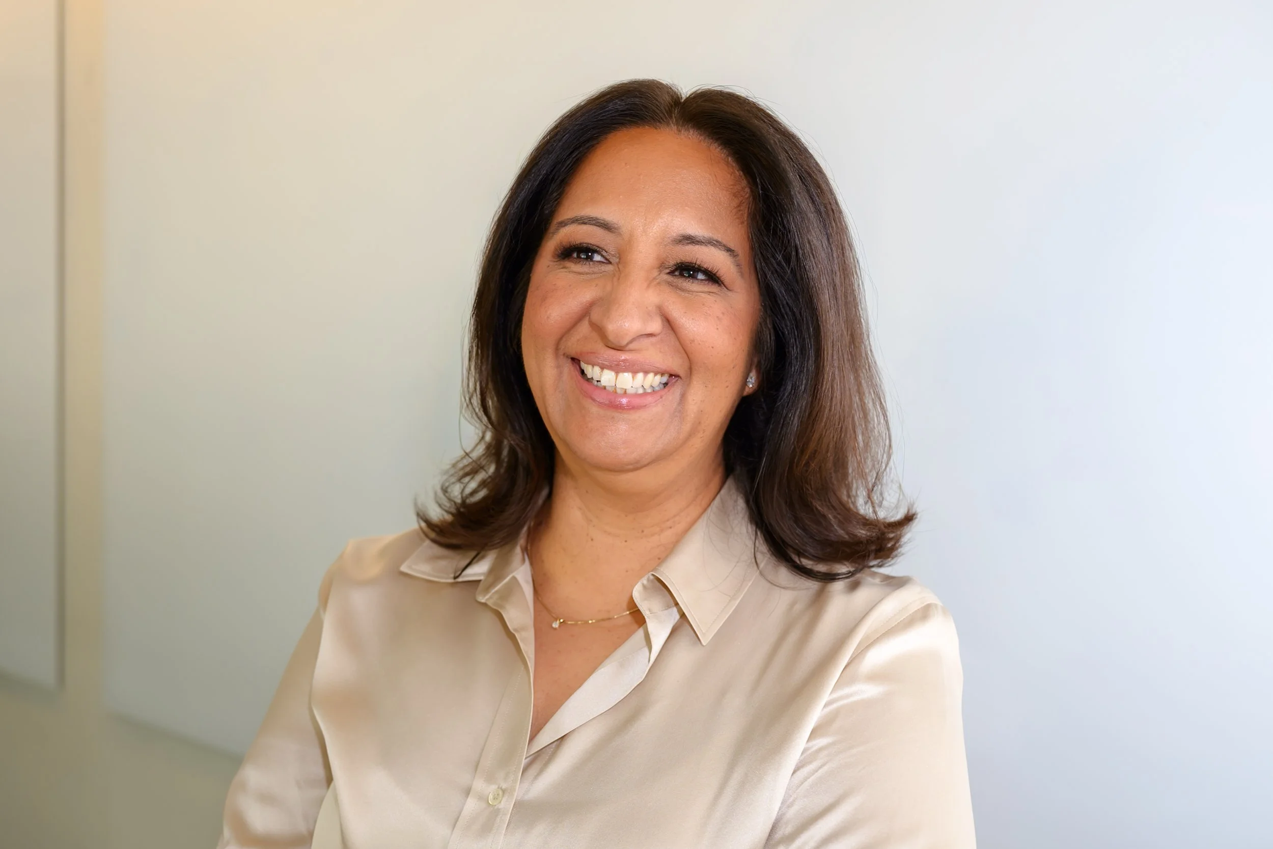 A woman with shoulder-length brown hair, smiling, wearing a beige button-up shirt and a delicate necklace, standing against a plain light-colored wall.