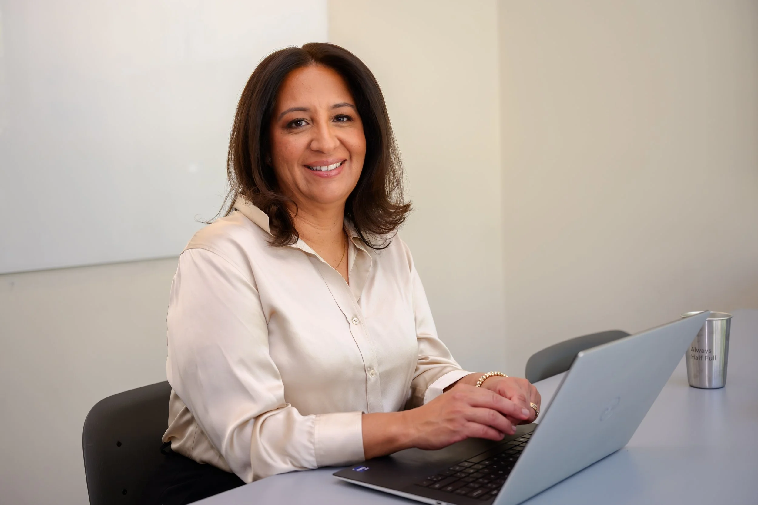 A woman with shoulder-length dark brown hair, wearing a cream-colored blouse, is sitting at a table and working on a silver laptop in a plain, light-colored room. A metallic tumbler with the phrase "Always Half Full" is on the table nearby.