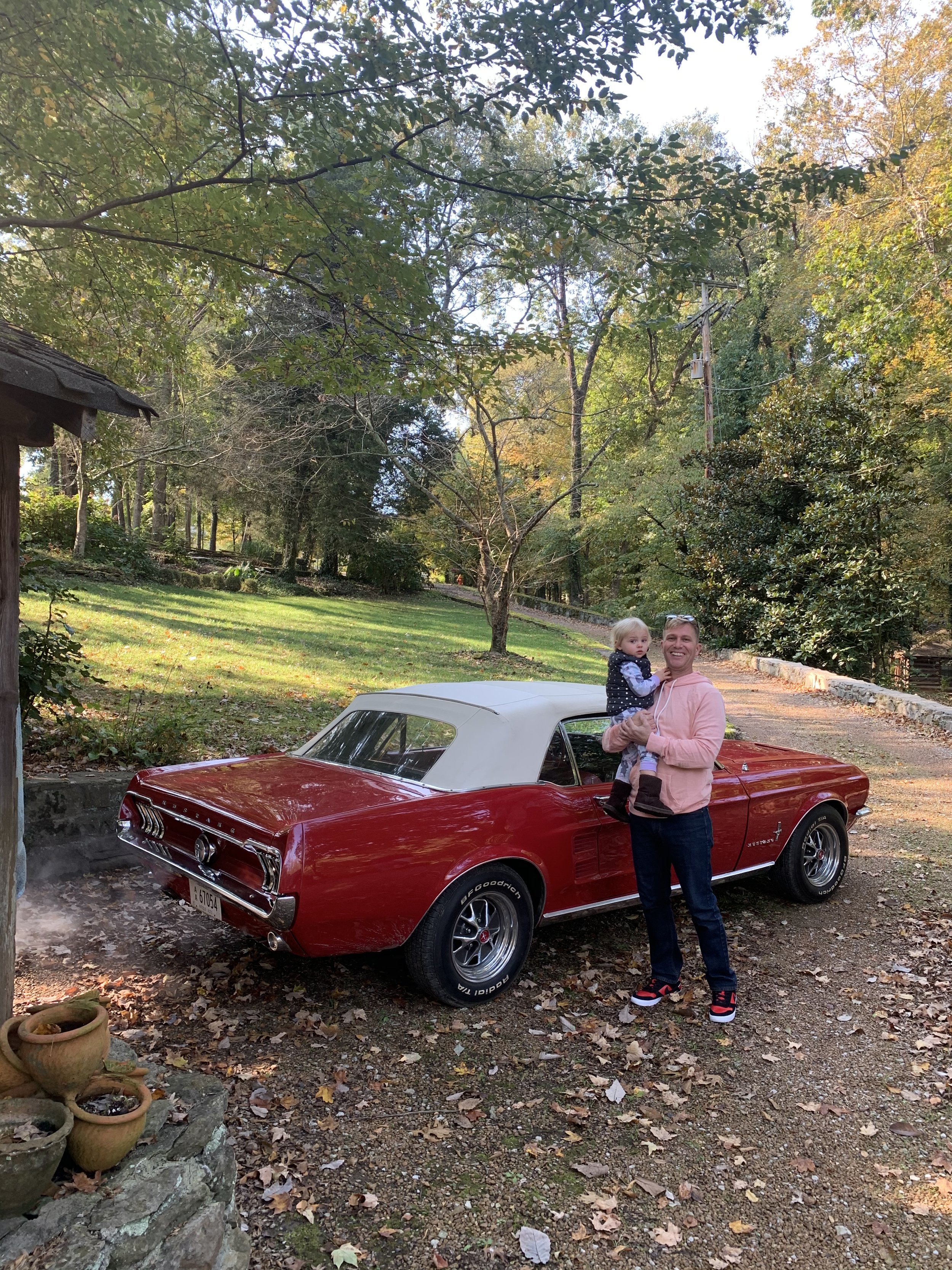 A man holding a young girl in front of a red vintage convertible car with a white top, parked on a wooded area with trees and fallen leaves.