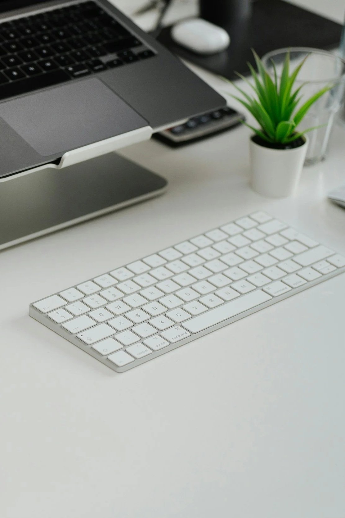 A white wireless keyboard on a white desk with a small potted green plant, a laptop, and a glass of water in the background.