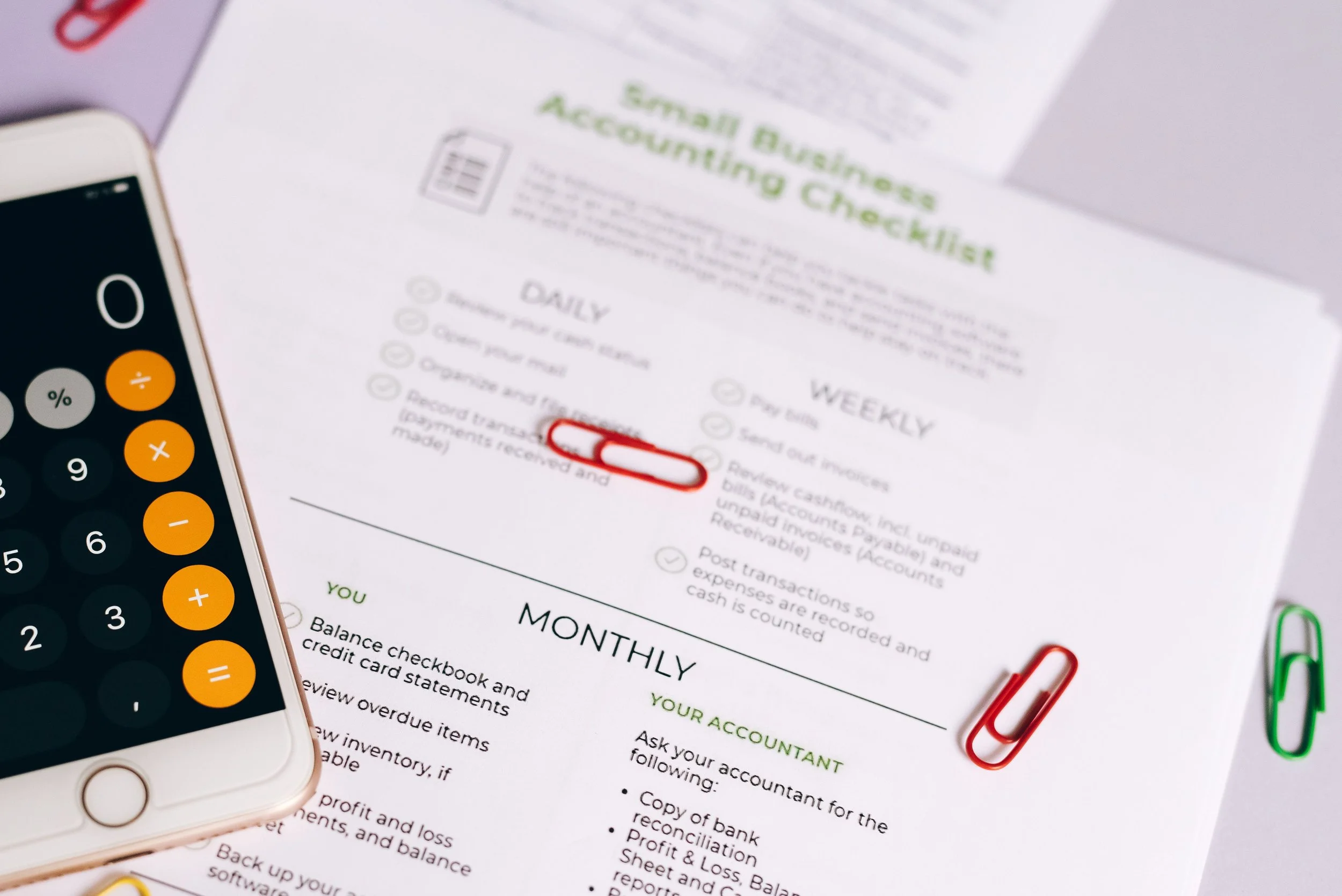 A printed sheet titled 'Small Business Accounting Checklist' with sections for Daily, Weekly, Monthly, and Yearly tasks. The sheet is next to a calculator and paper clips, with a smartphone partially visible in the foreground.