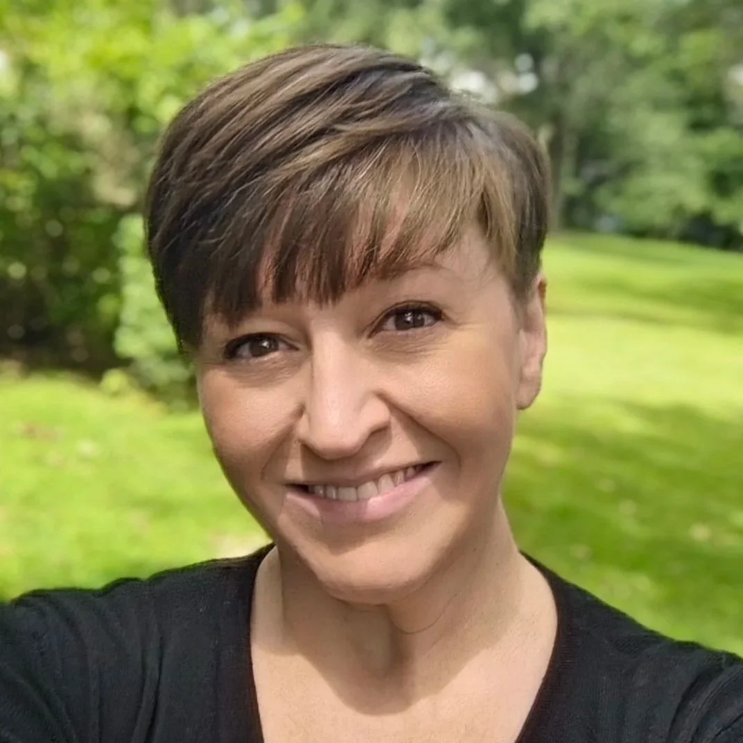 A woman with short brown hair and a black top smiling outdoors with a green, wooded background.
