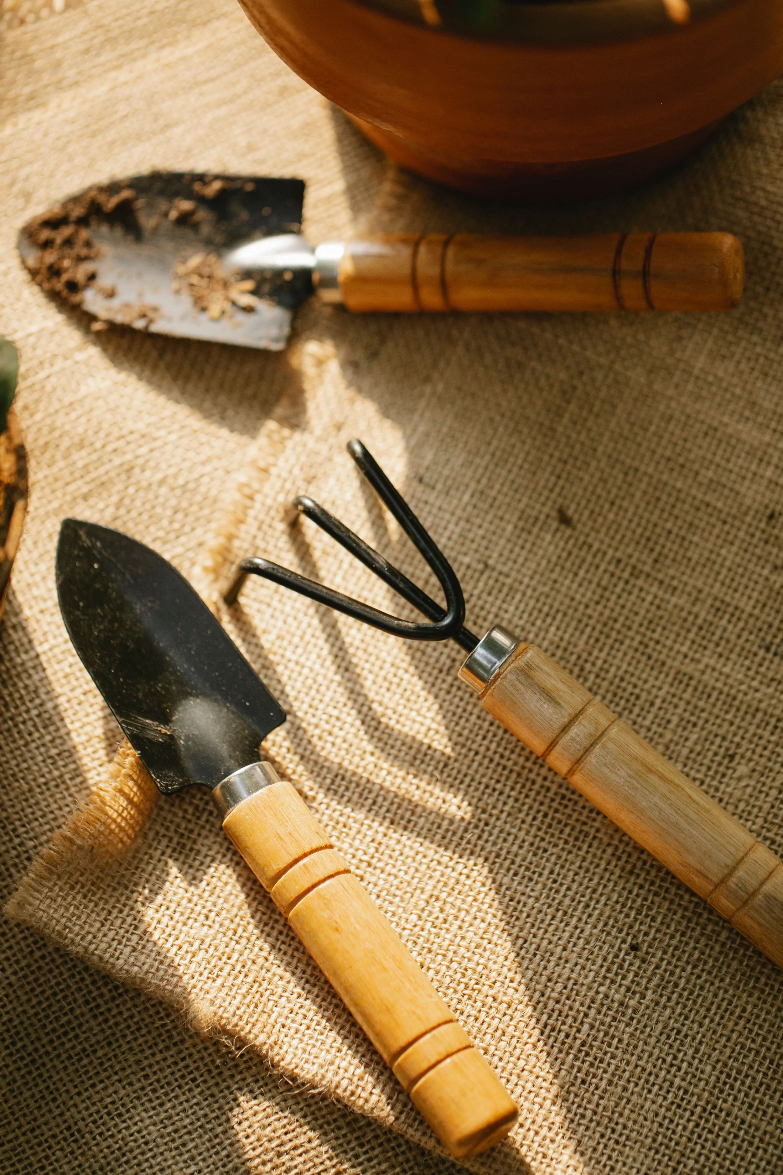 A set of gardening tools on yellowish burlap fabric, including a trowel with a black blade, a hand rake, and two other hand tools with wooden handles.