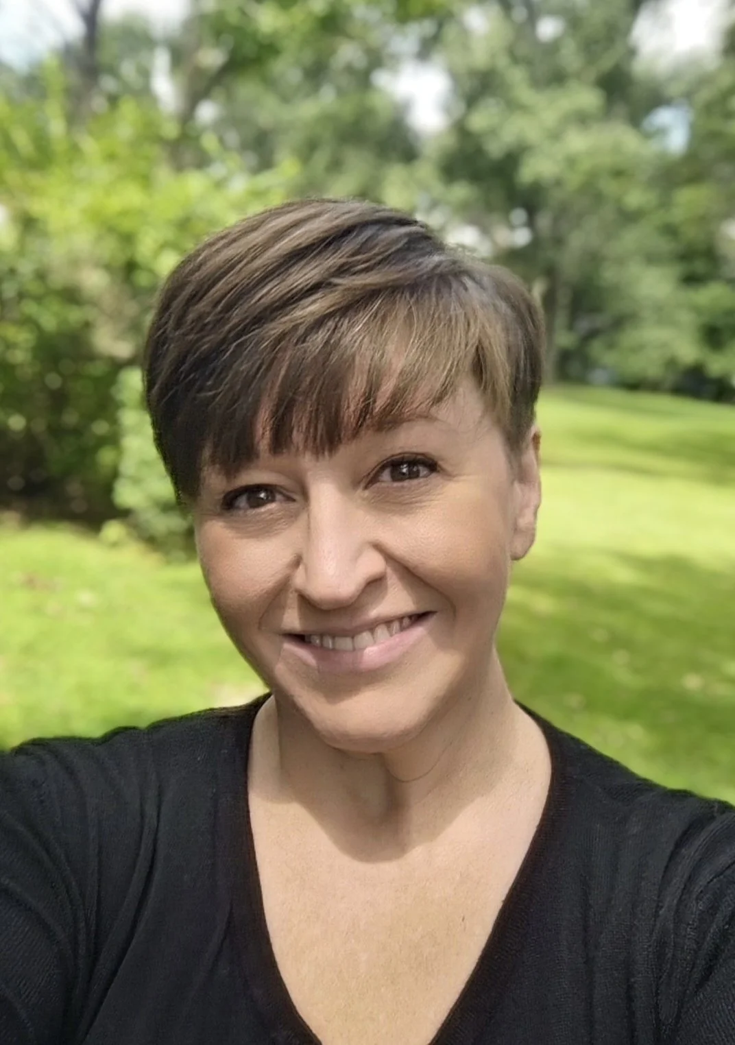 Smiling woman with short brown hair taking a selfie outdoors, with green trees and grass in the background.