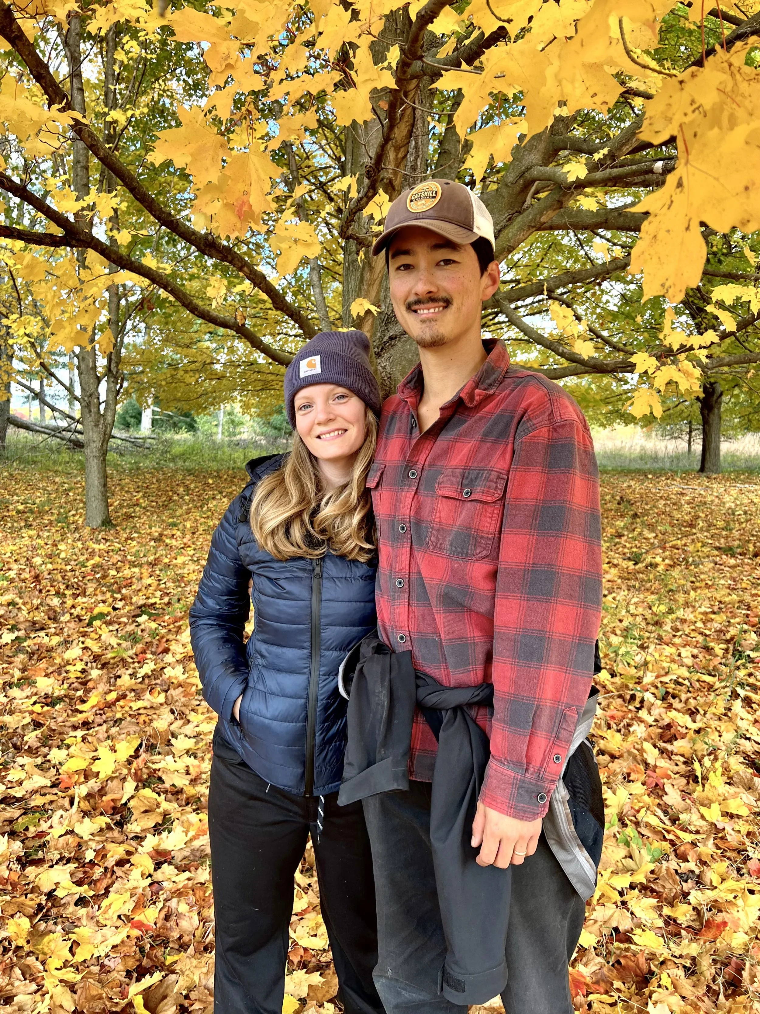 A man and a woman are standing outdoors in an autumnal park with a tree behind them full of yellow and orange leaves. They are smiling at the camera. The man is wearing a brown cap with a logo and a red plaid shirt, while the woman is wearing a dark beanie with a logo and a blue jacket. The ground is covered with fallen yellow leaves.