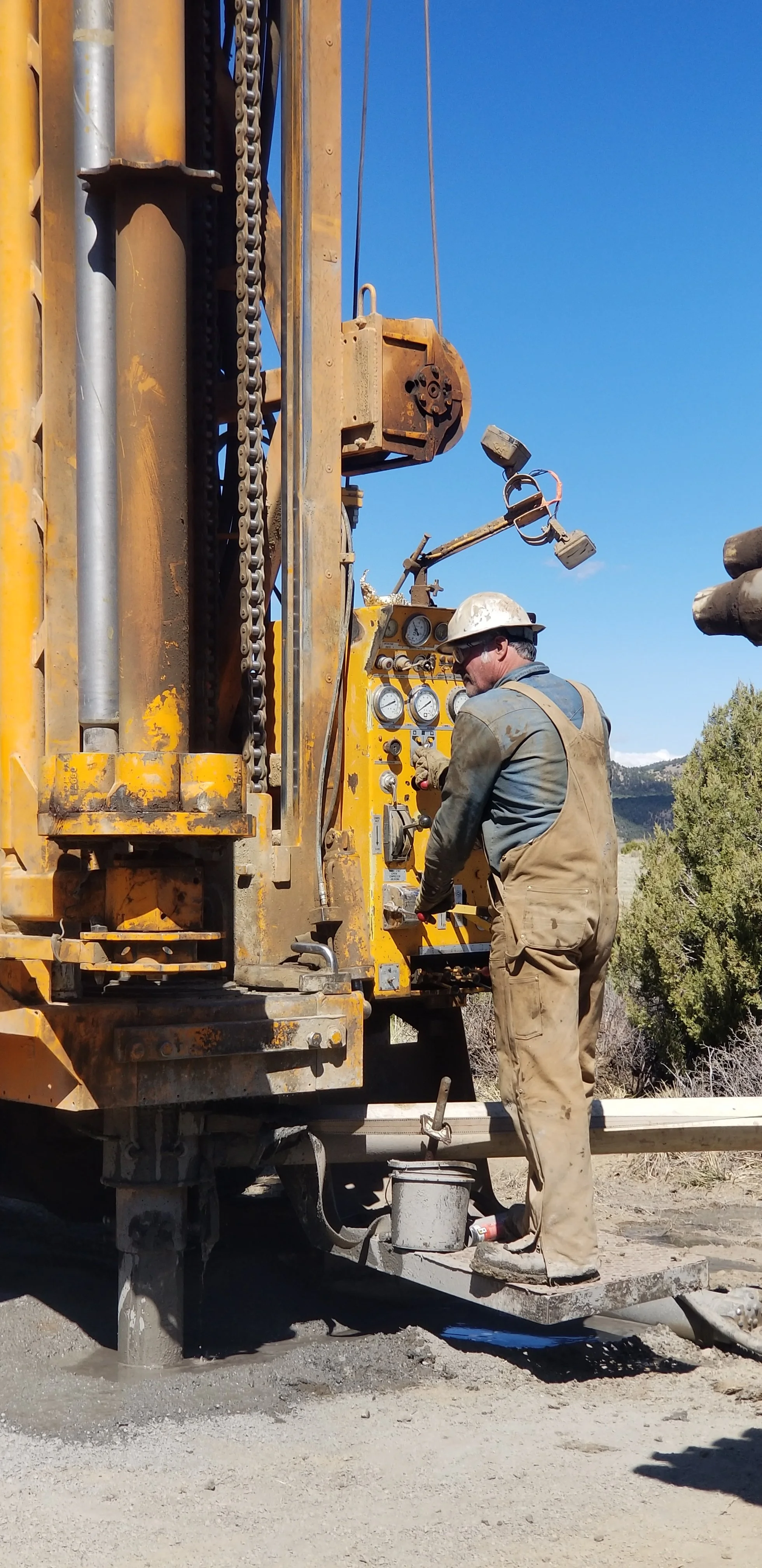 Brett Verney operating the Veryney Well Drilling rig on a gravel road in a rural area, with trees and mountains in the background under a clear blue sky.