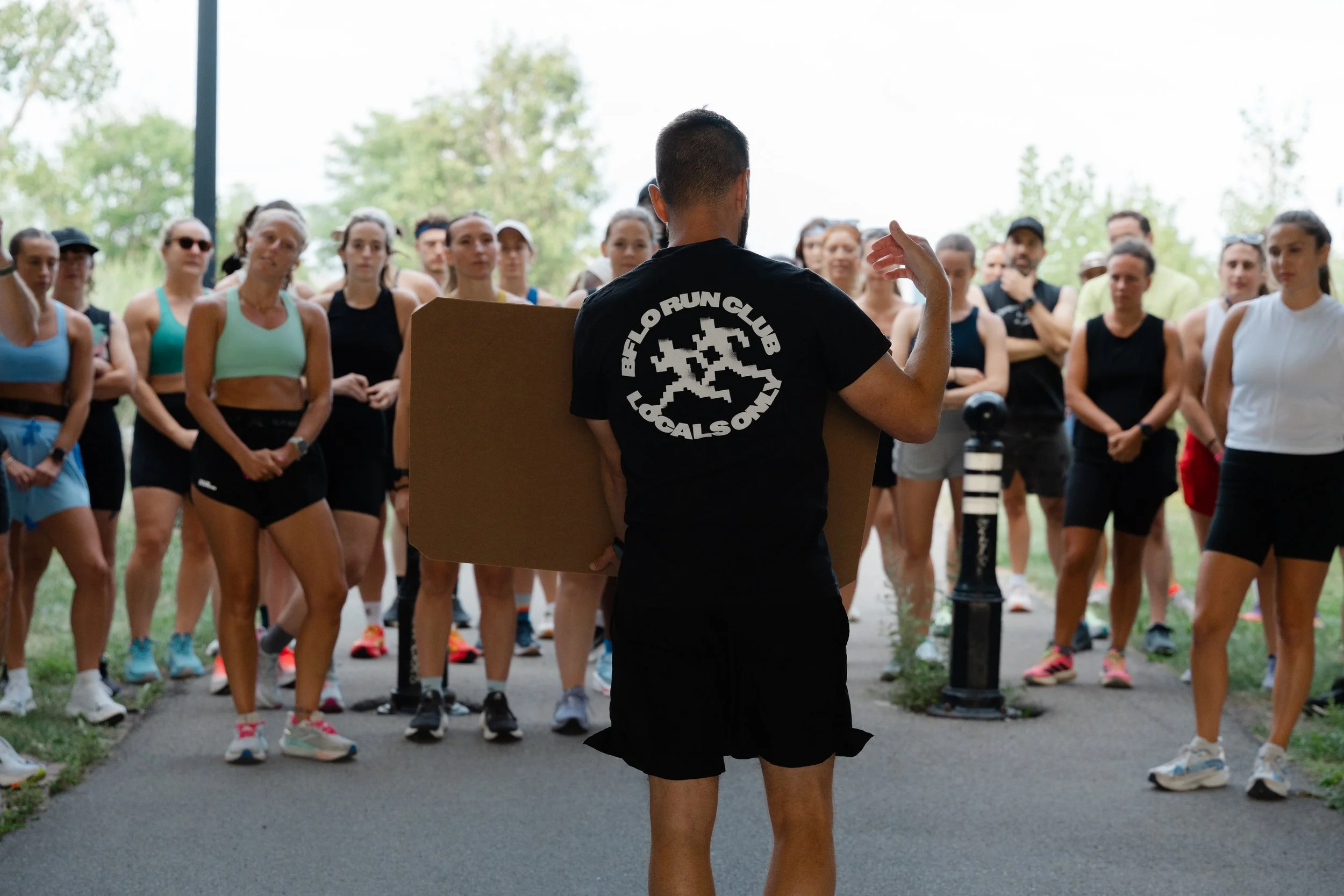 A man in a black shirt with a logo on the back is speaking to a group of women and a few men in athletic clothing outdoors during the daytime, in a park or similar setting.