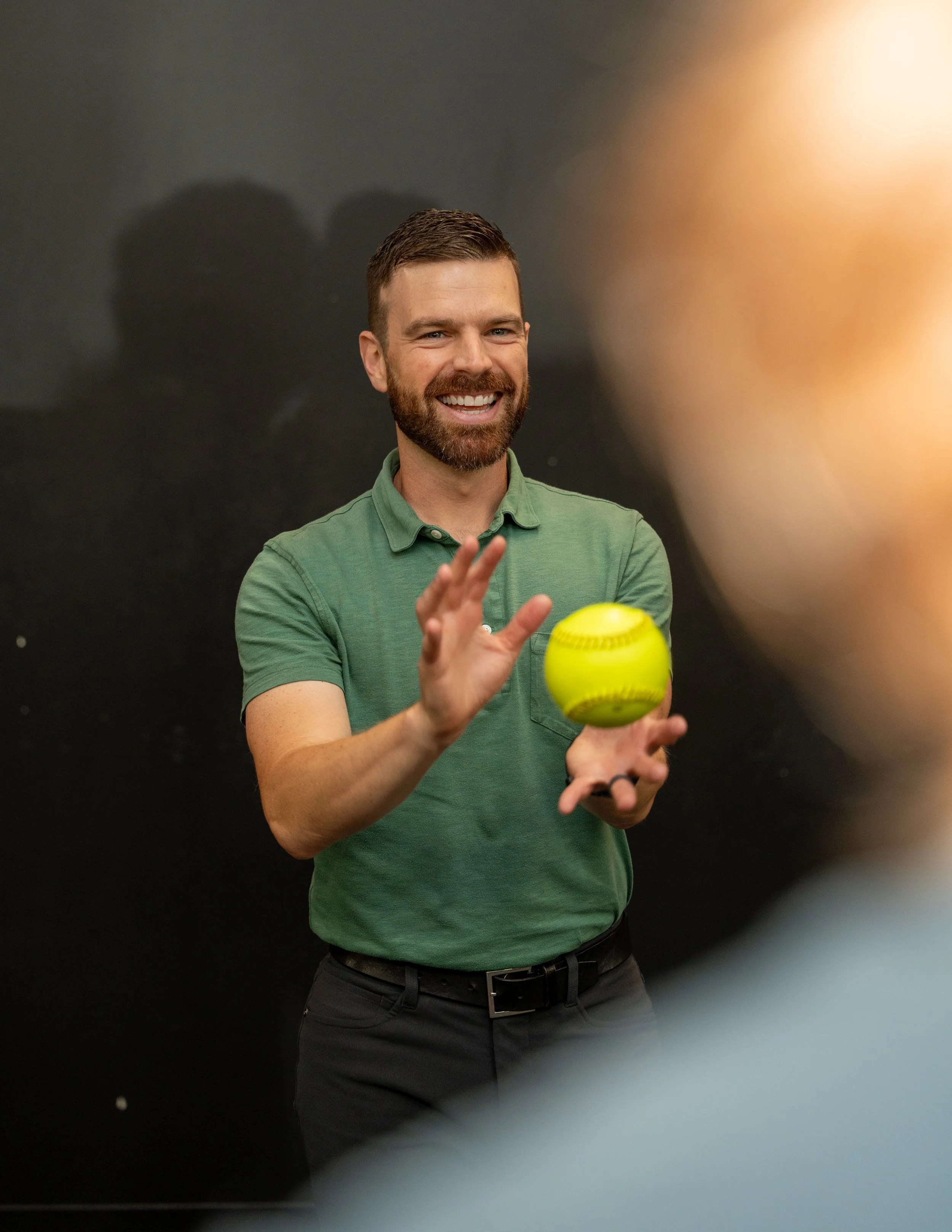 A man with a beard and short brown hair smiling and holding a yellow tennis ball, standing against a dark background.