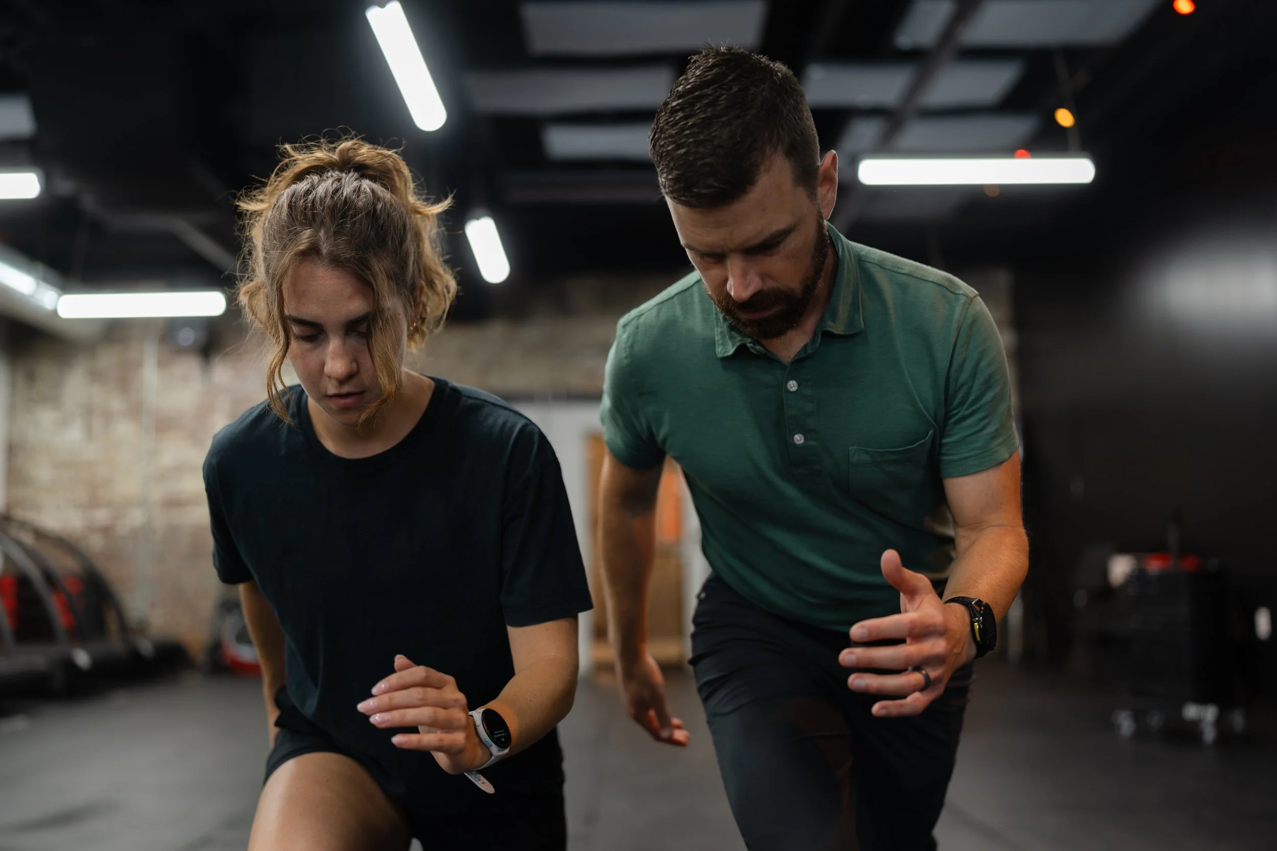 Two people, a woman and a man, exercising together indoors in a gym. They are bent over with their hands on their knees, engaging in a stretching or warm-up activity. The gym has modern lighting and exposed brick walls.