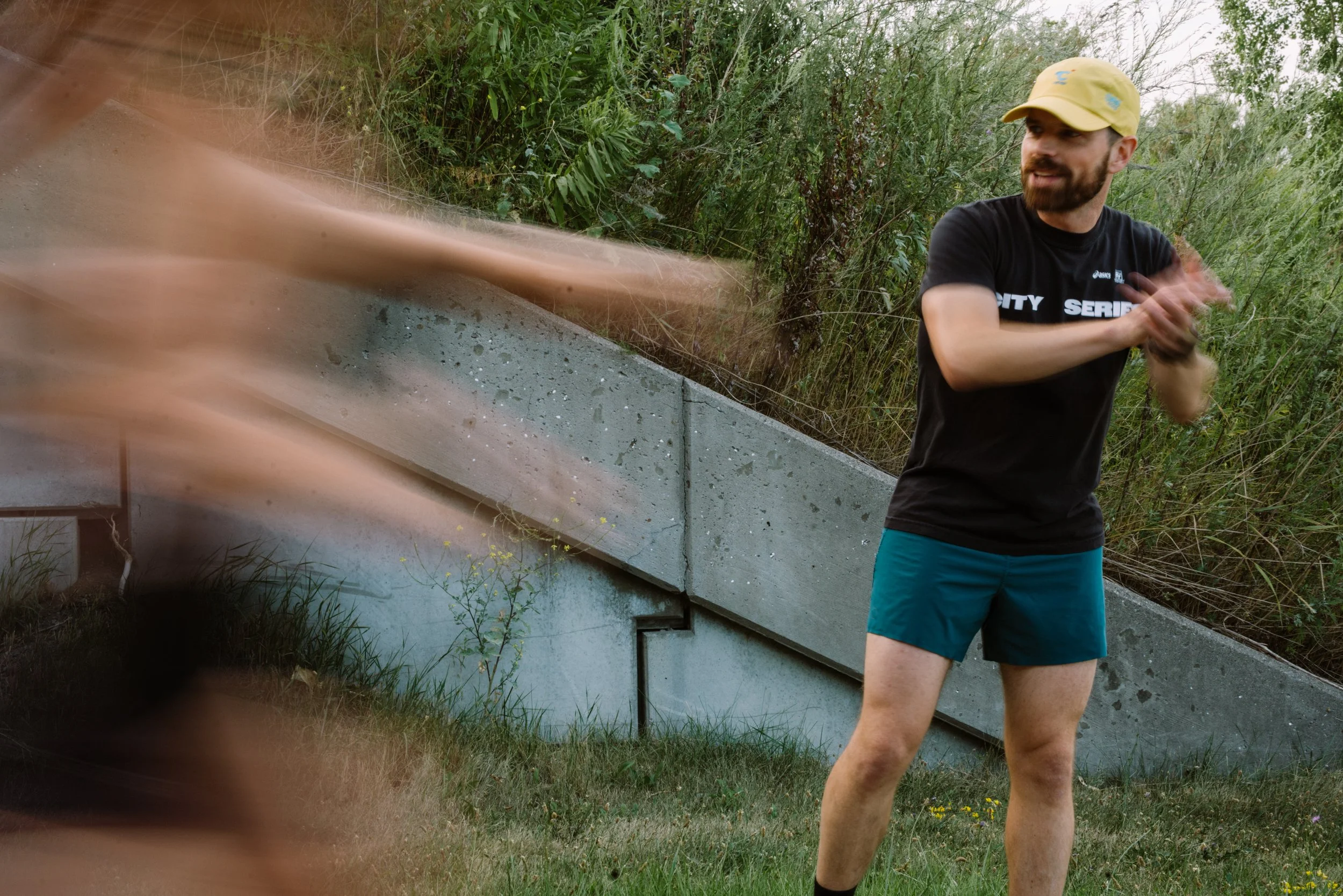 A man with a beard in a black shirt and teal shorts standing outside near bushes, clapping, wearing a yellow cap, and with a concrete wall in the background.