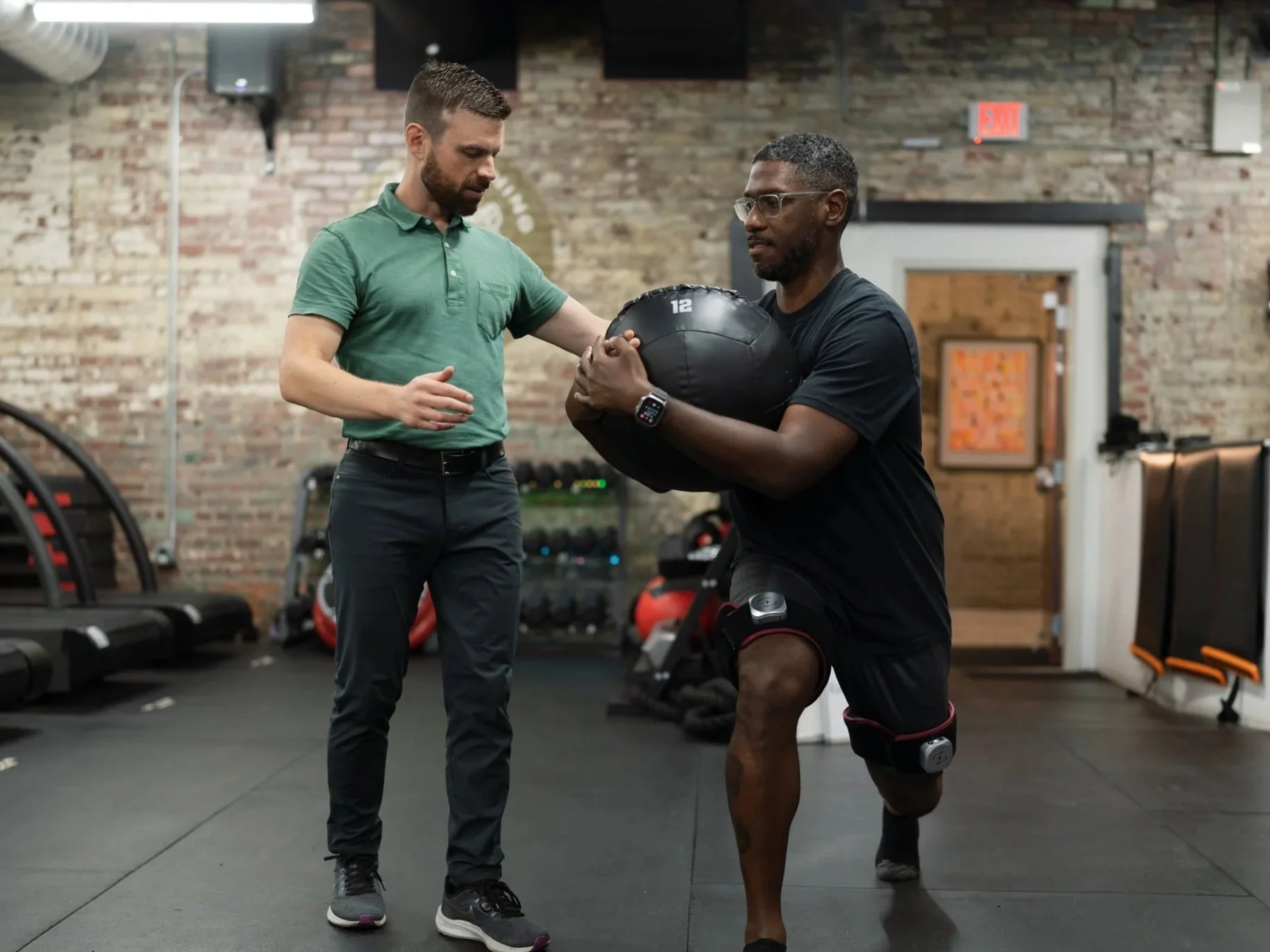 Two men in a gym, one guiding the other on how to perform a physical exercise with a medicine ball, with brick walls and workout equipment in the background.