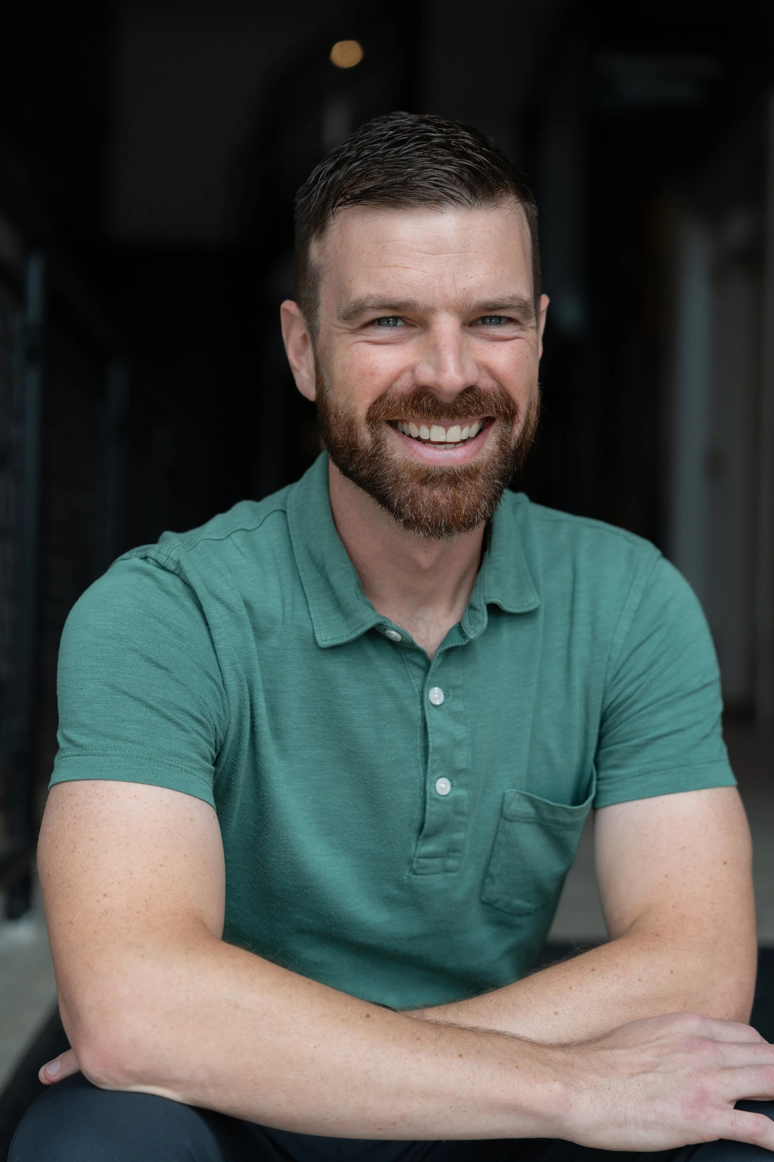 A smiling man with a beard and short brown hair, wearing a green polo shirt, sitting with his arms crossed at a table, in a dark indoor setting.