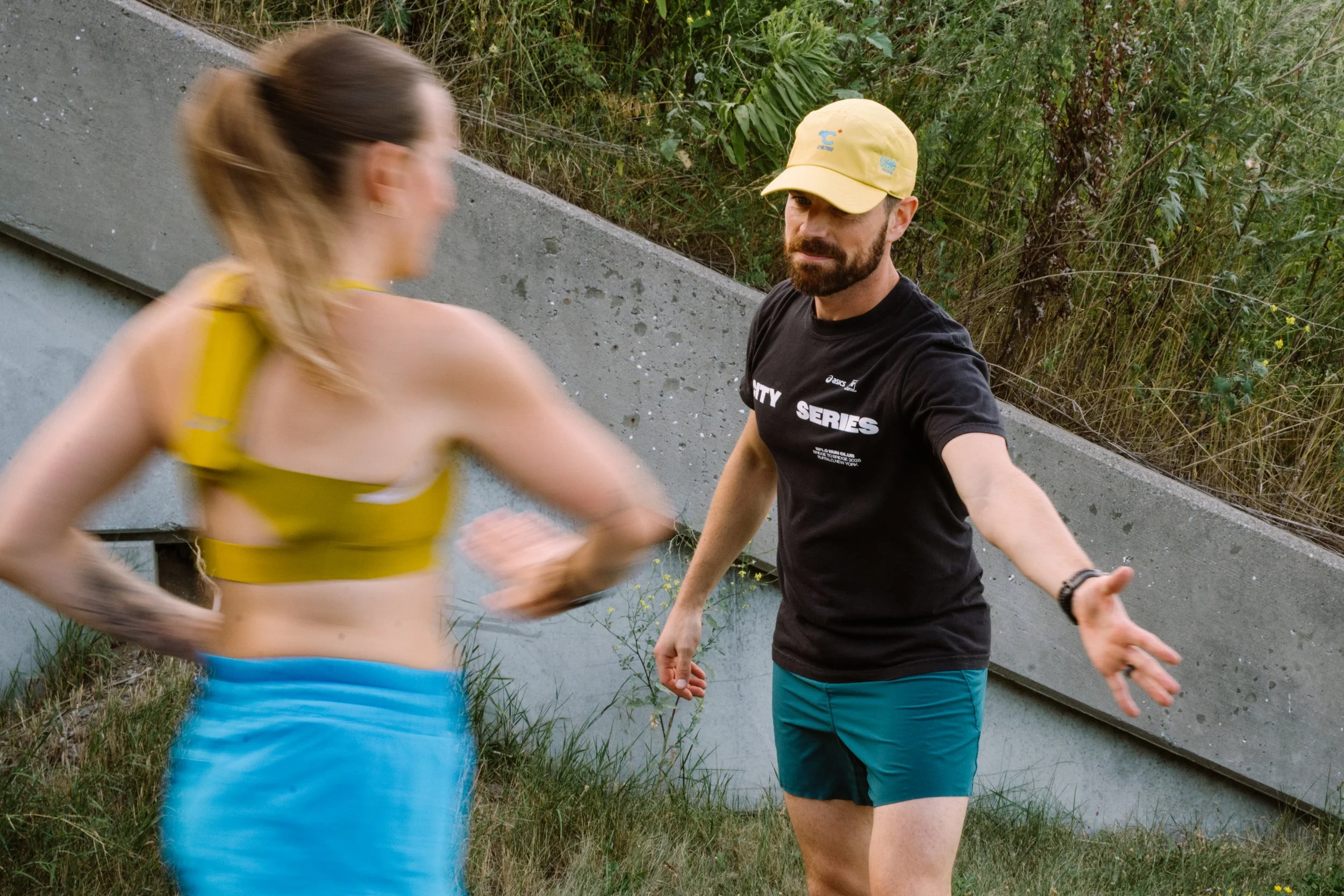 A man and woman running outdoors on a concrete path surrounded by greenery. The woman is in the foreground, wearing a yellow sports bra and bright blue leggings, with motion blur. The man, in the background, has a beard, wearing a black T-shirt, teal shorts, and a tan cap, looking at the woman.