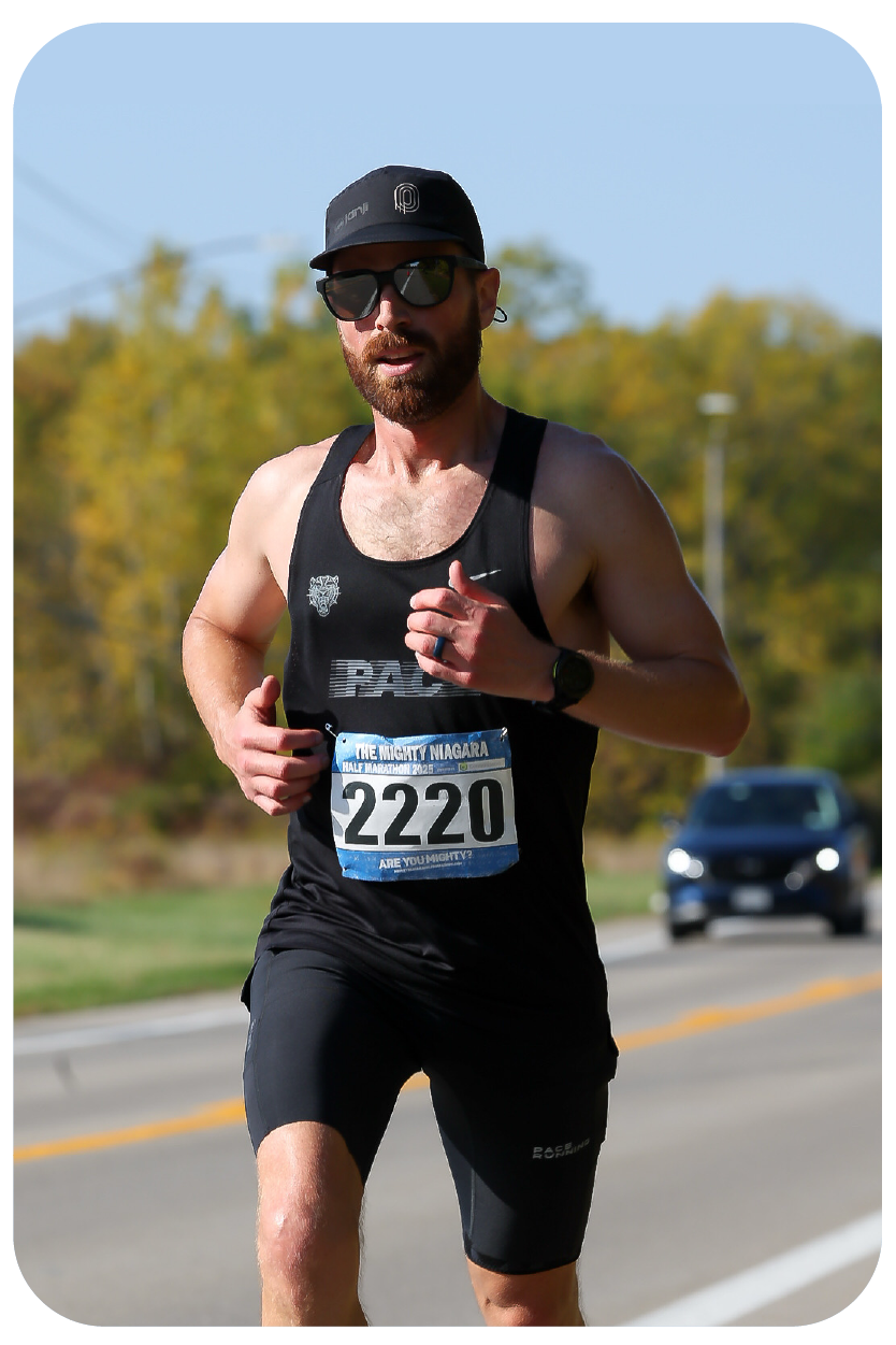 A man running in a marathon, wearing a black sleeveless athletic shirt, black shorts, a black cap, sunglasses, and a racing bib number 2220, on a scenic road with trees and a car in the background.