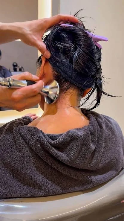 Person receiving a scalp micropigmentation tattoo treatment on the back of their head, with a technician using a tattoo machine, while the person has hair clips and a towel around their neck.