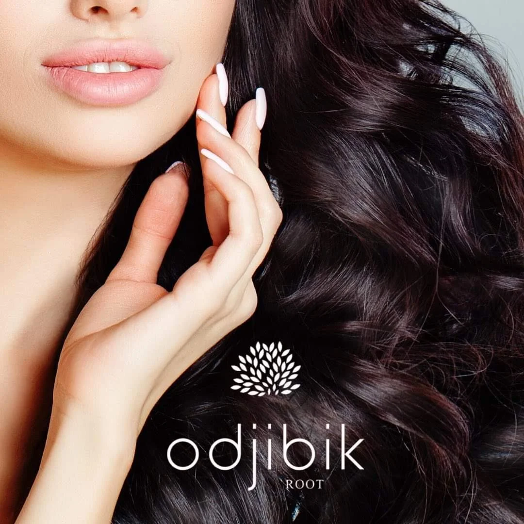 Close-up of a woman with long, dark, curly hair, lightly colored lips, and manicured nails touching her hair. The logo 'odjibik ROOT' is in the lower part of the image.