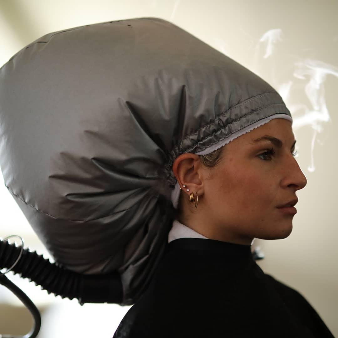 A woman with multiple earrings, wearing a black turtleneck, has a large gray hood over her head, likely a hydrodermabrasion or facial treatment device, with steam rising in the background.