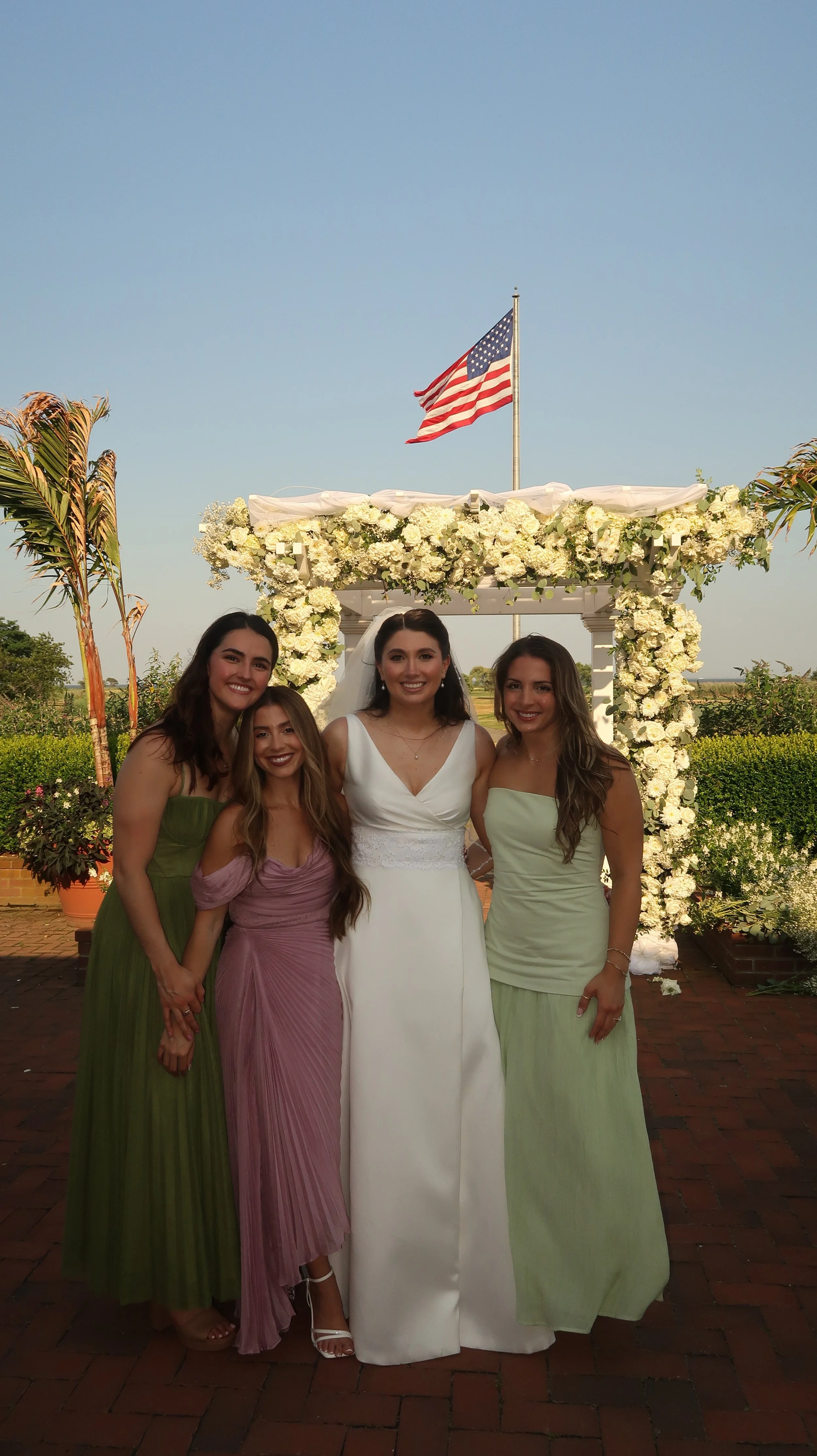 Five women standing together in front of a floral arch with an American flag flying above, outdoors on a sunny day.