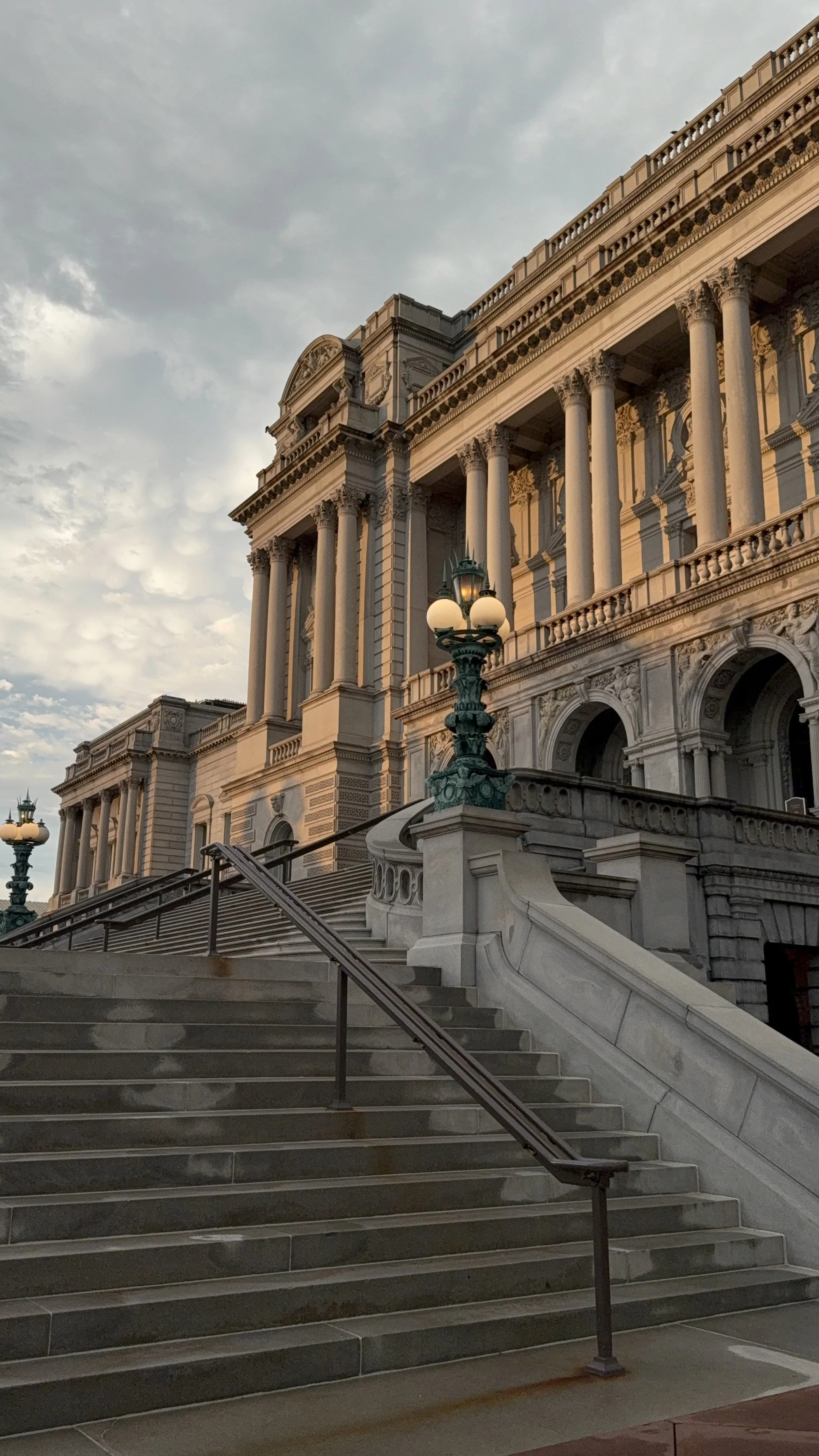 Exterior of a grand historic building with classical architecture, including columns and ornate details, with stone stairs and vintage street lamps, under a cloudy sky at dusk.