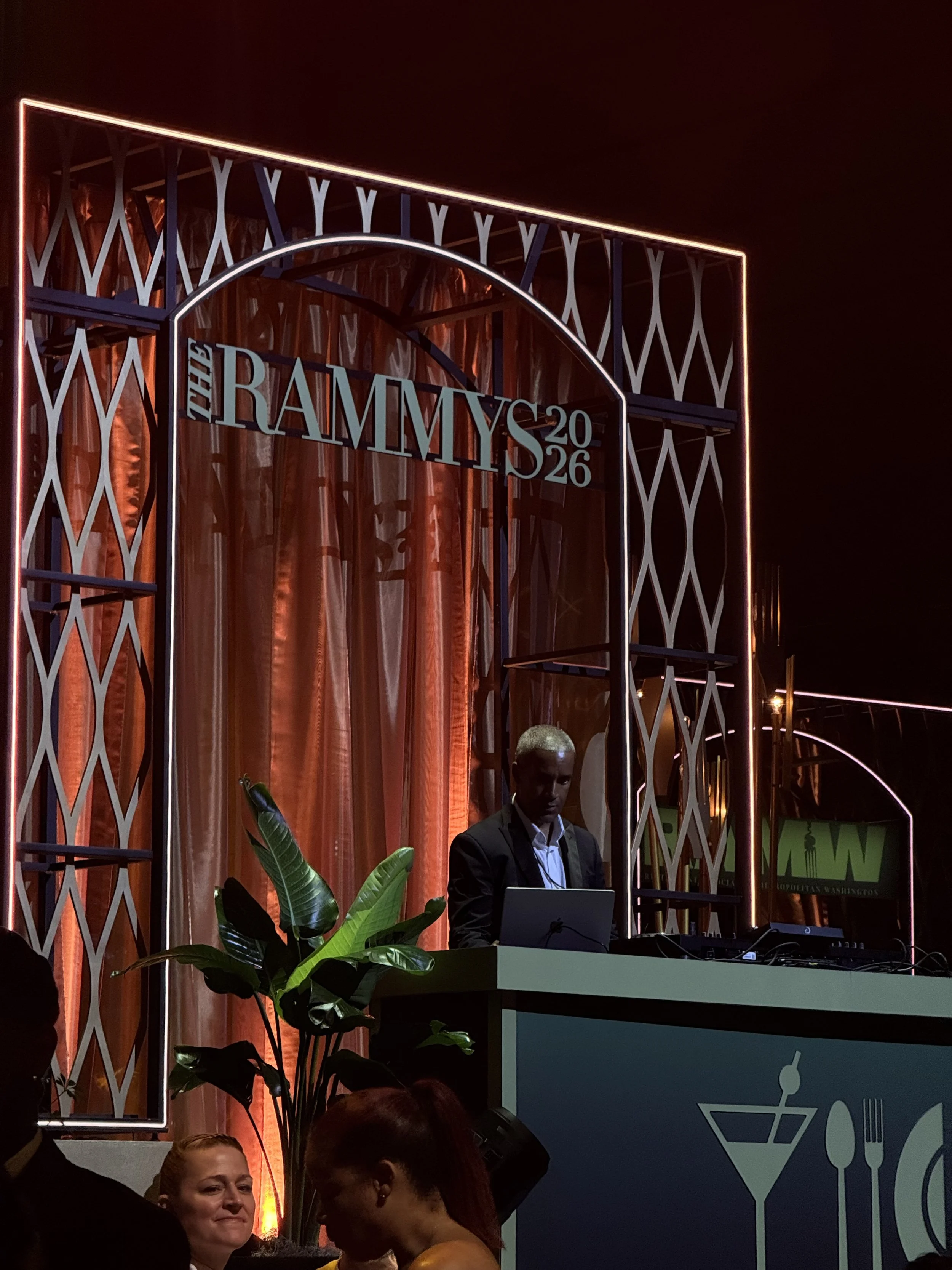 Night scene at an event with a stage, illuminated sign reading 'The Rammys 2026', man using DJ equipment, decorative backdrop, plants, multiple people in foreground, and illuminated accents on structures.