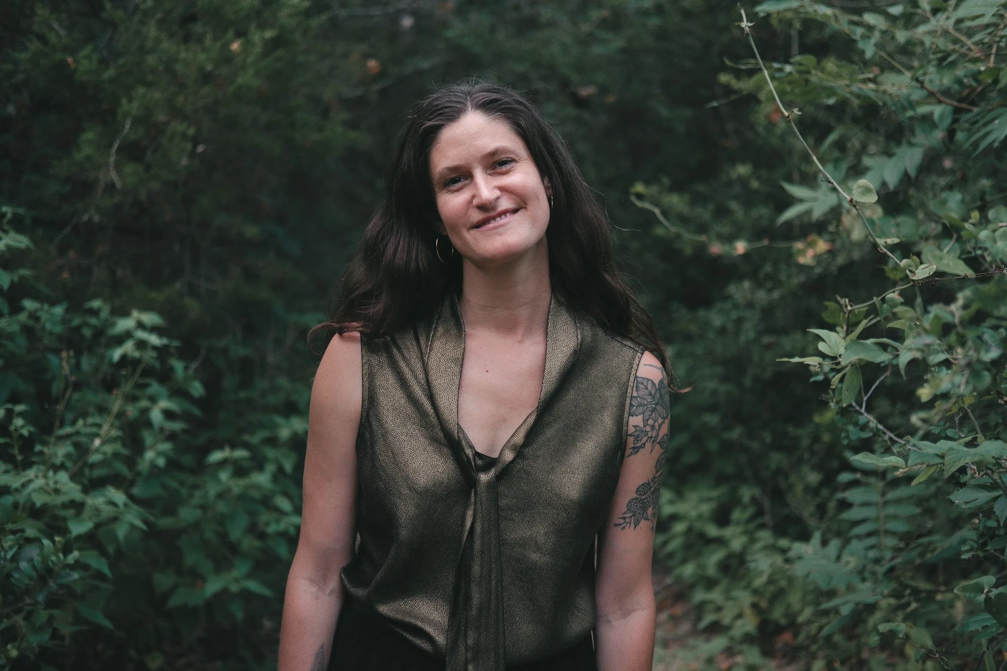A woman with dark wavy hair and a tattoo on her right arm smiling outdoors in front of green foliage.