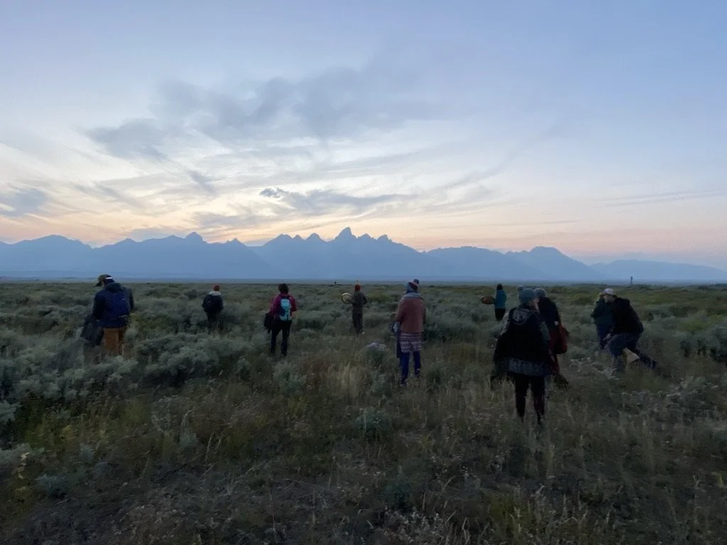 Group of people walking across a grassy plain at sunset with mountains in the background.