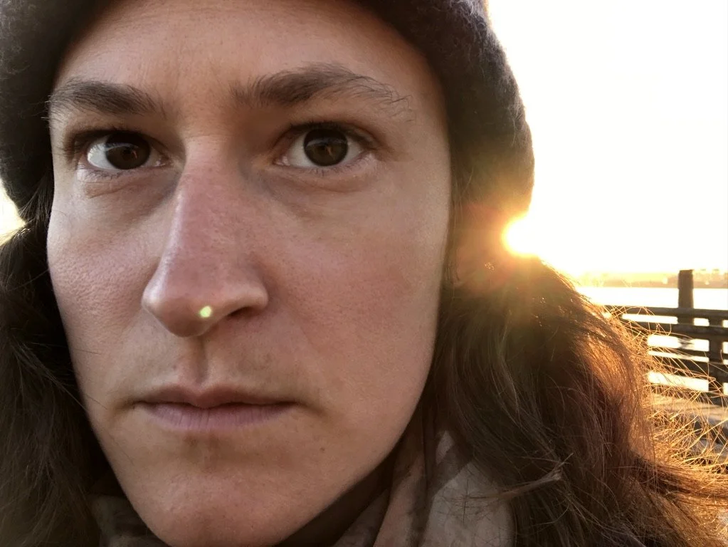 Close-up of a woman's face with brown eyes and long, wavy brown hair, outdoors during sunrise.