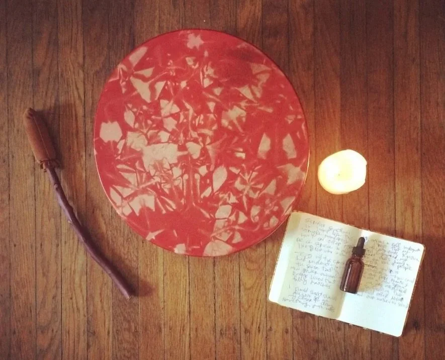 A wooden floor with a red drum,  lit candle, and an open notebook with handwritten notes and a small brown glass dropper bottle.