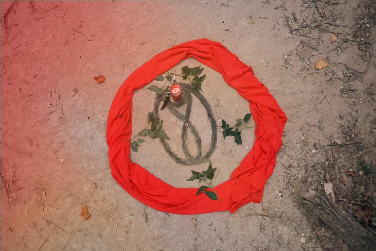 A ceremonial display on the ground with a red cloth arranged in a circle, green leaves, a lit candle, and a drawn black sigil (abstract symbol).