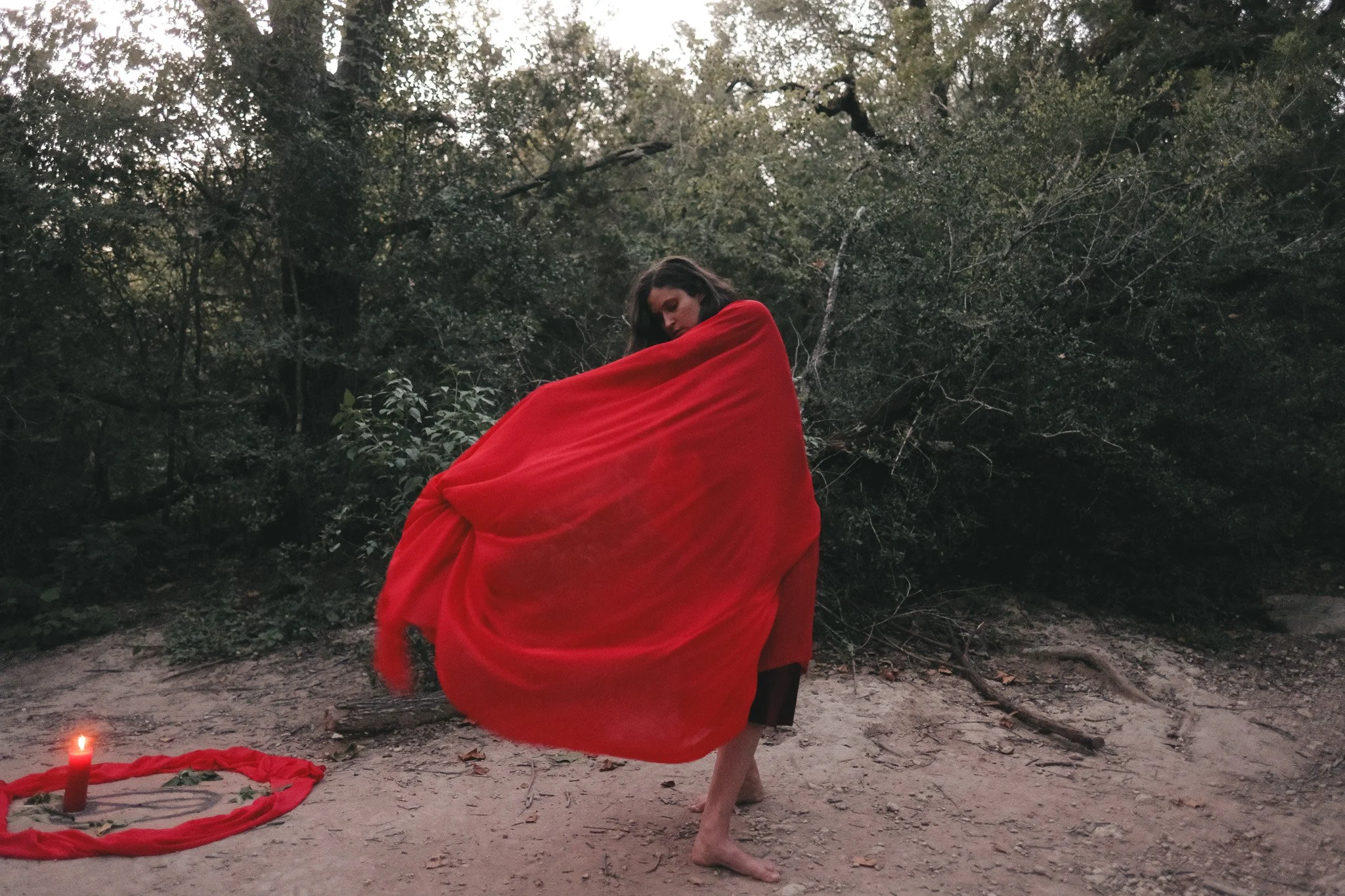 A woman dressed in a flowing red fabric stands barefoot outdoors in a natural setting with trees and dirt ground. There is a red candle and cloth on the ground nearby.