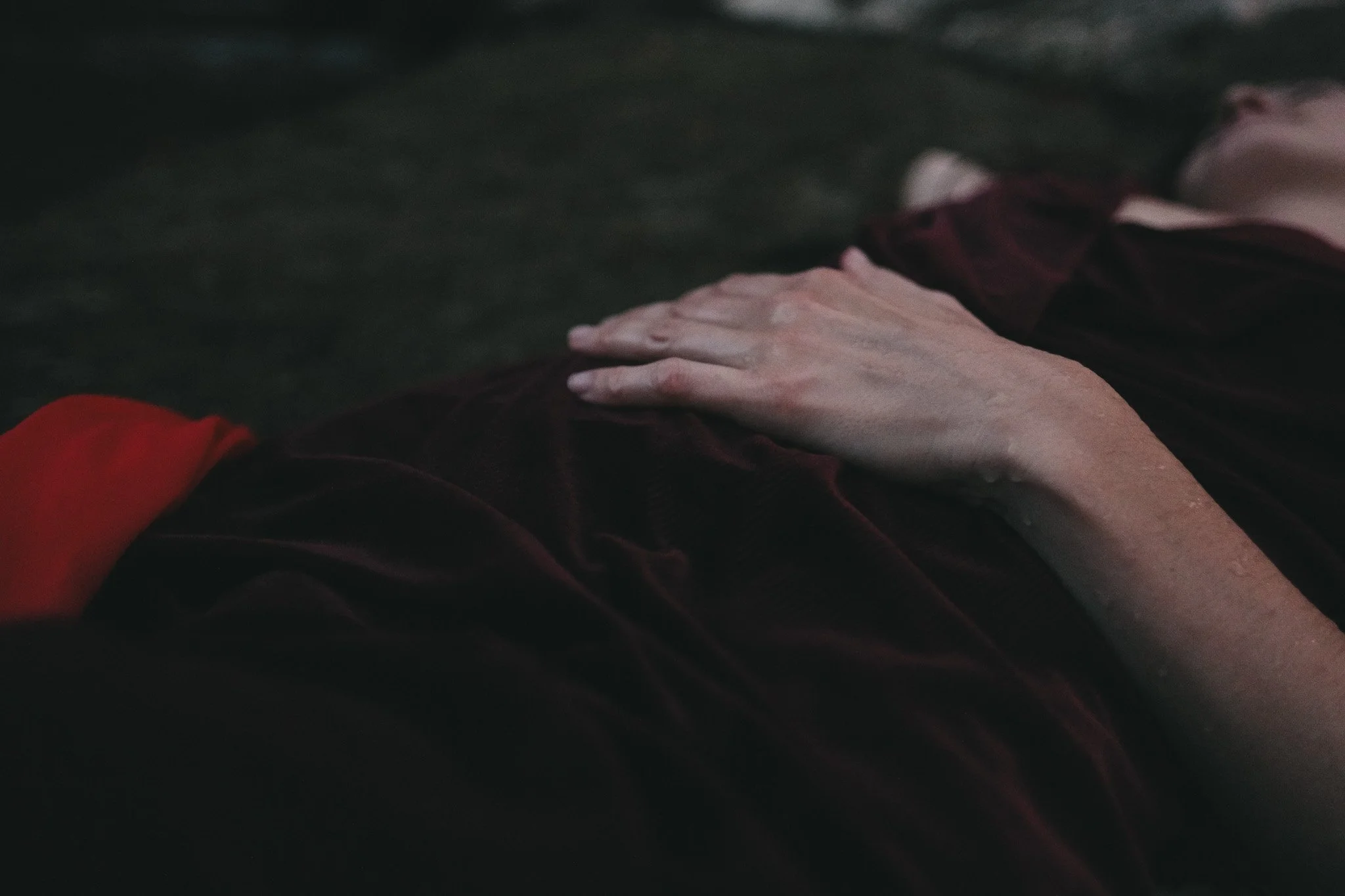 Woman in dark velvet dress laying in a stream with one hand resting on her belly and a red fabric draped over her leg.