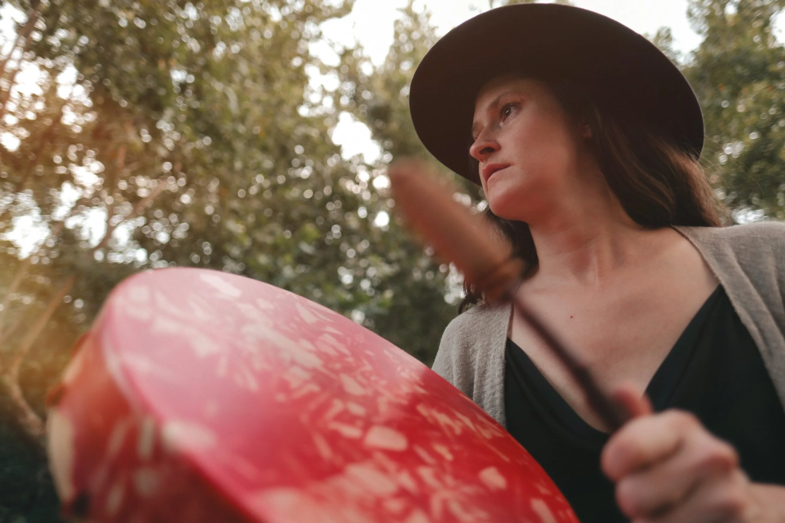A woman wearing a black hat playing a red drum and looking into the distance outdoors with trees in the background.