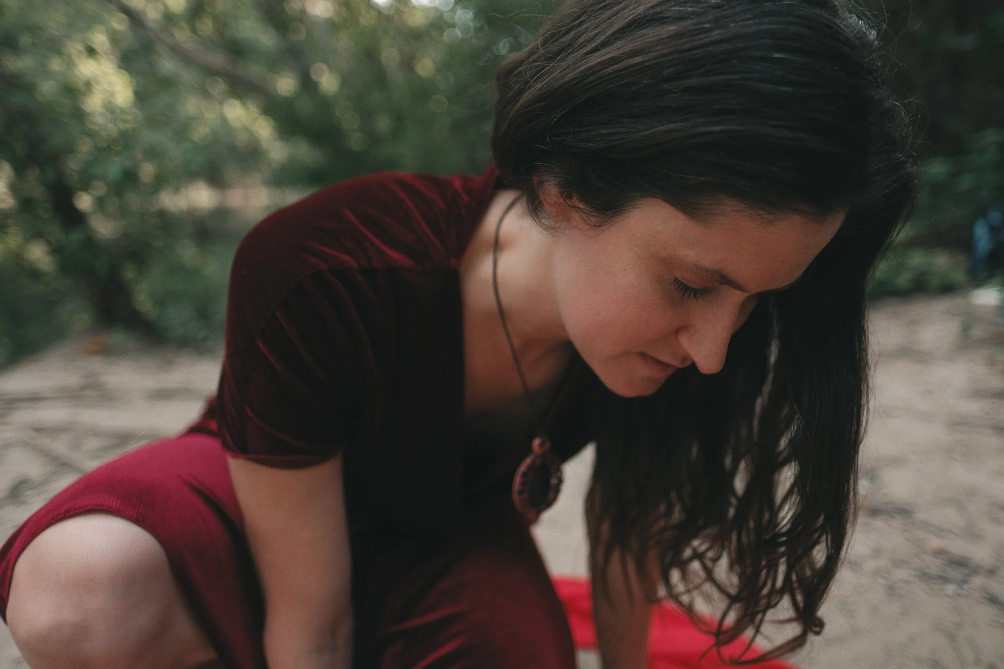A woman with long dark hair, wearing a dark red dress, is squatting and looking down at the ground with her eyes closed with trees in the background.