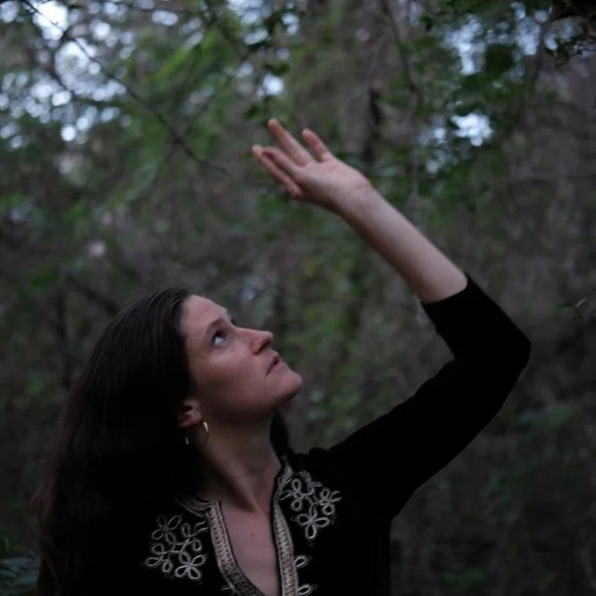 A woman with dark hair and earrings reaching up towards the branches of trees in a wooded area.