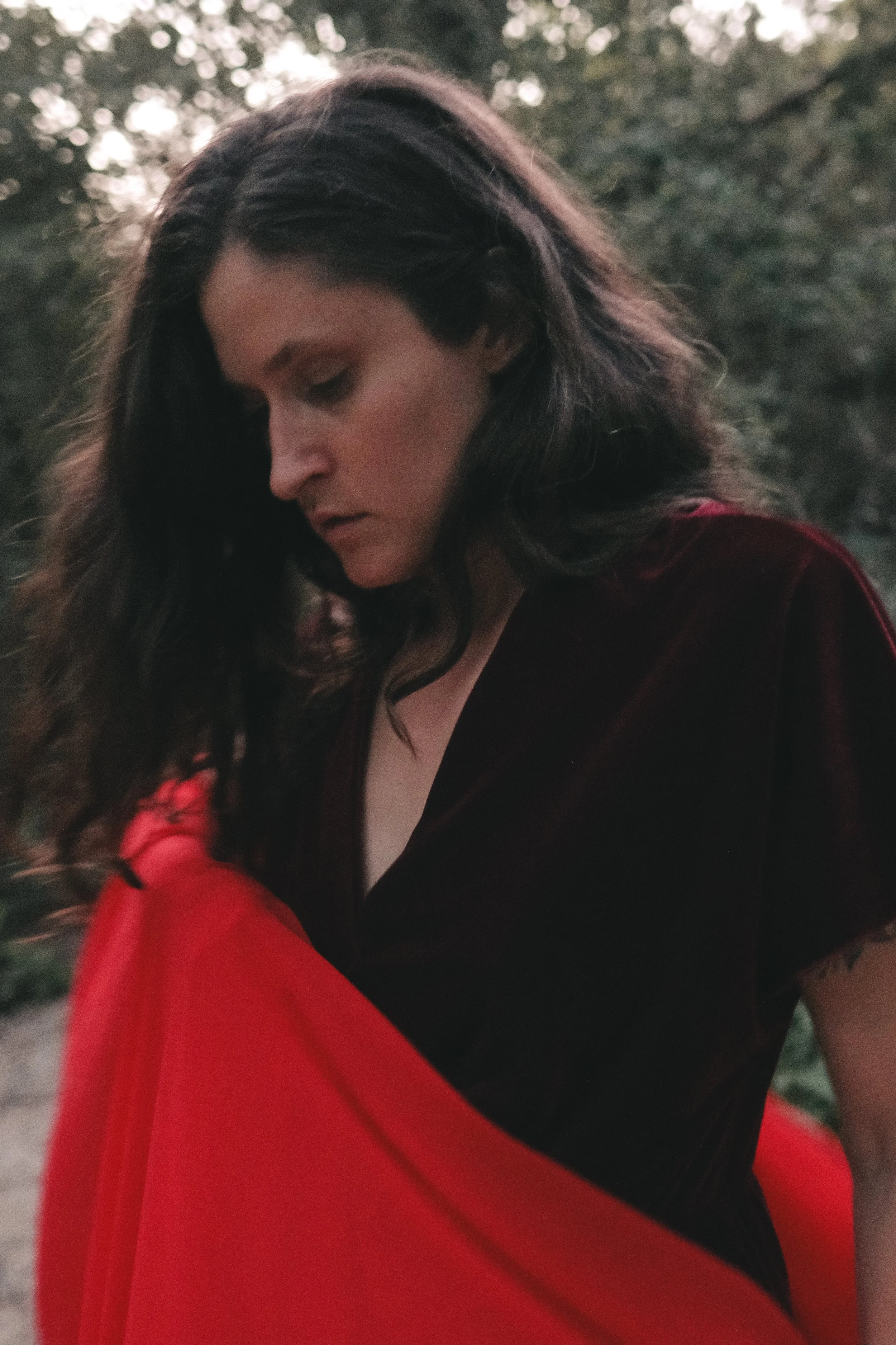 A woman with dark, wavy hair looks down, wearing a dark dress top and wrapping a red skirt fabric around her, standing outdoors with blurred trees and sky in the background.