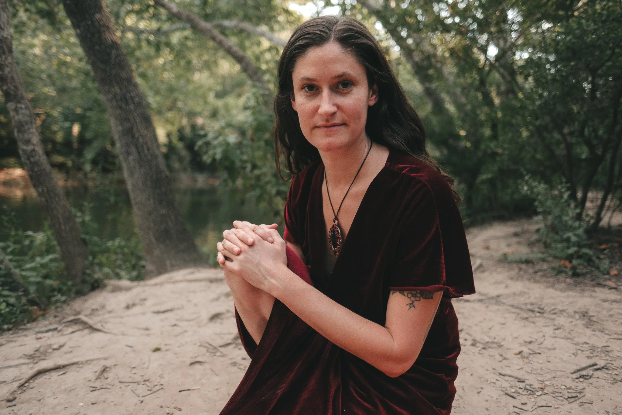 A woman with long dark hair and tattoos on her arms, wearing a dark red velvet dress, kneels with hands clasped in front of her in a wooded outdoor setting during daytime.